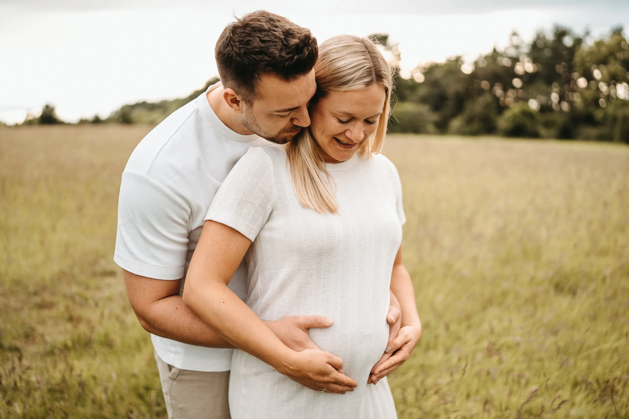 Ein Paar steht in einem Feld, der Mann umarmt die schwangere Frau from hinten und küsst sie auf die Wange, bei Sonnenuntergang.