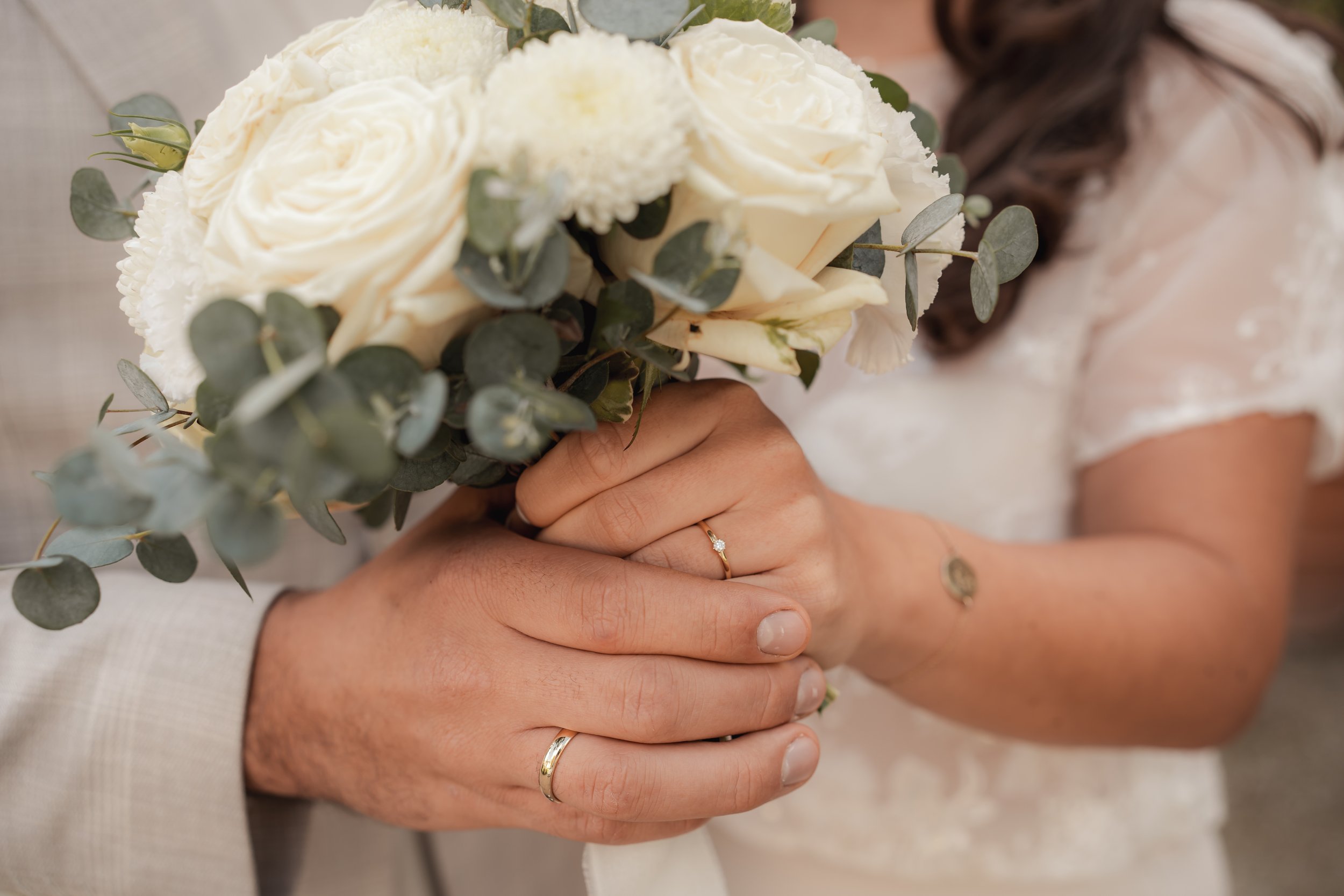 Hände eines Paares halten sich, beide tragen Eheringe. Die Frau hält einen Brautstrauß mit weißen Rosen und Eukalyptus. Die Braut trägt ein weißes Hochzeitskleid.
