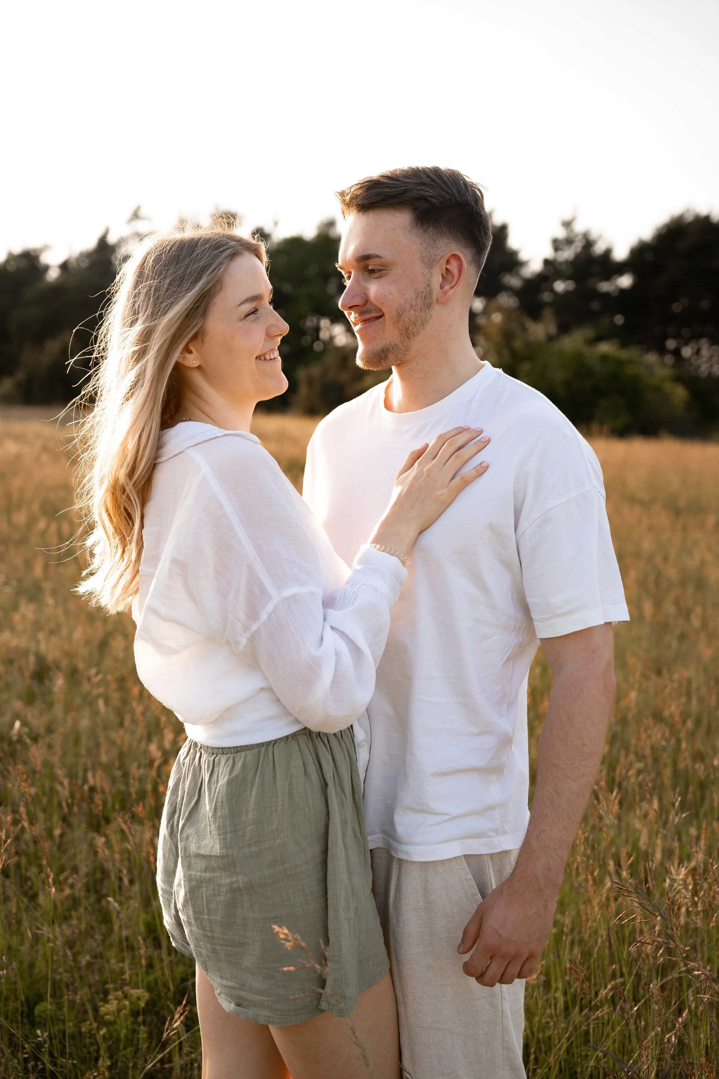 Ein junges Paar steht sich in einem Feld bei Sonnenuntergang gegenüber, lächelnd und sich liebevoll berührend.