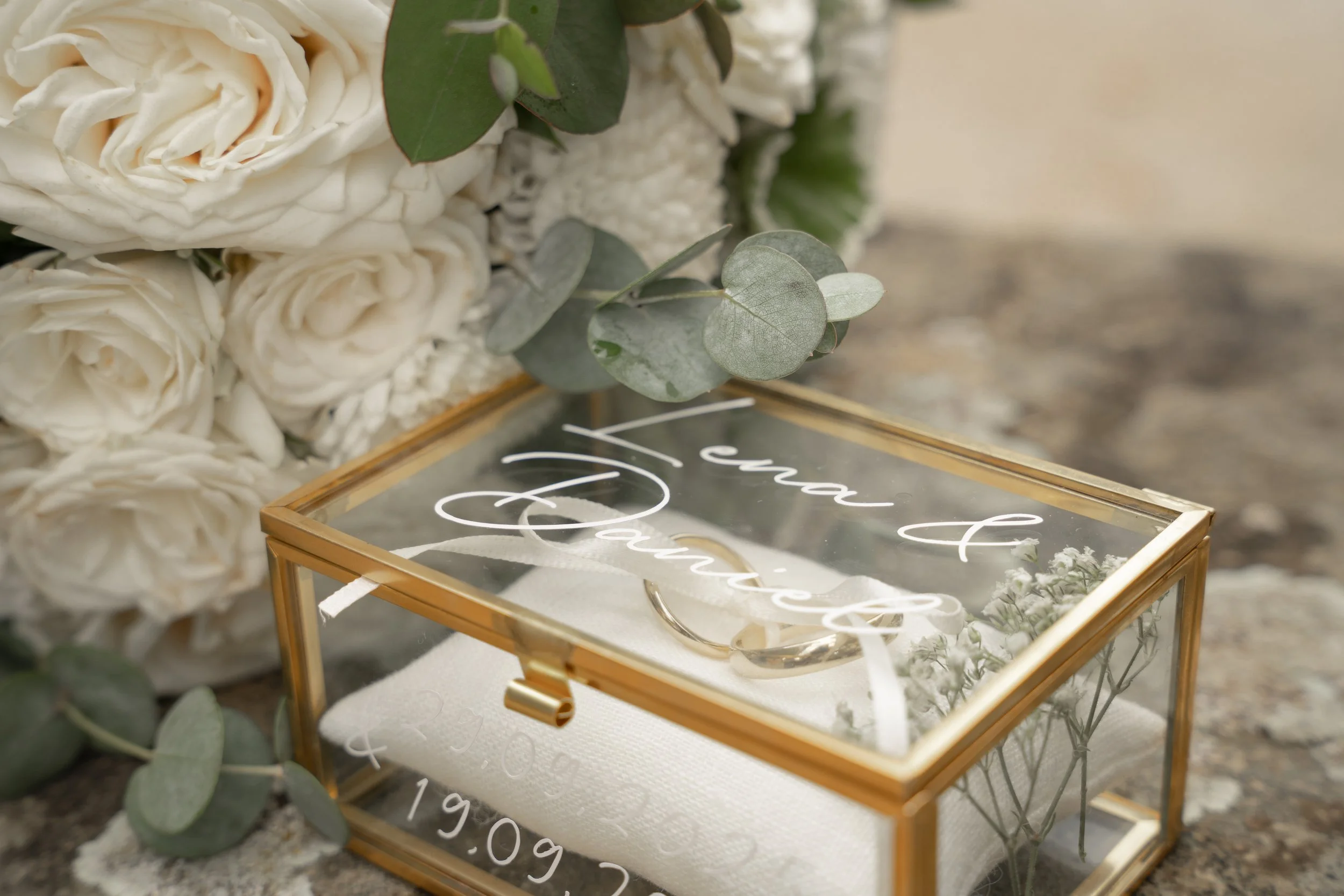Ein Hochzeitsring in einer goldfarbenen Glasbox mit Beschriftung 'love & forever', begleitet von weißen Rosen und grünen Blättern.