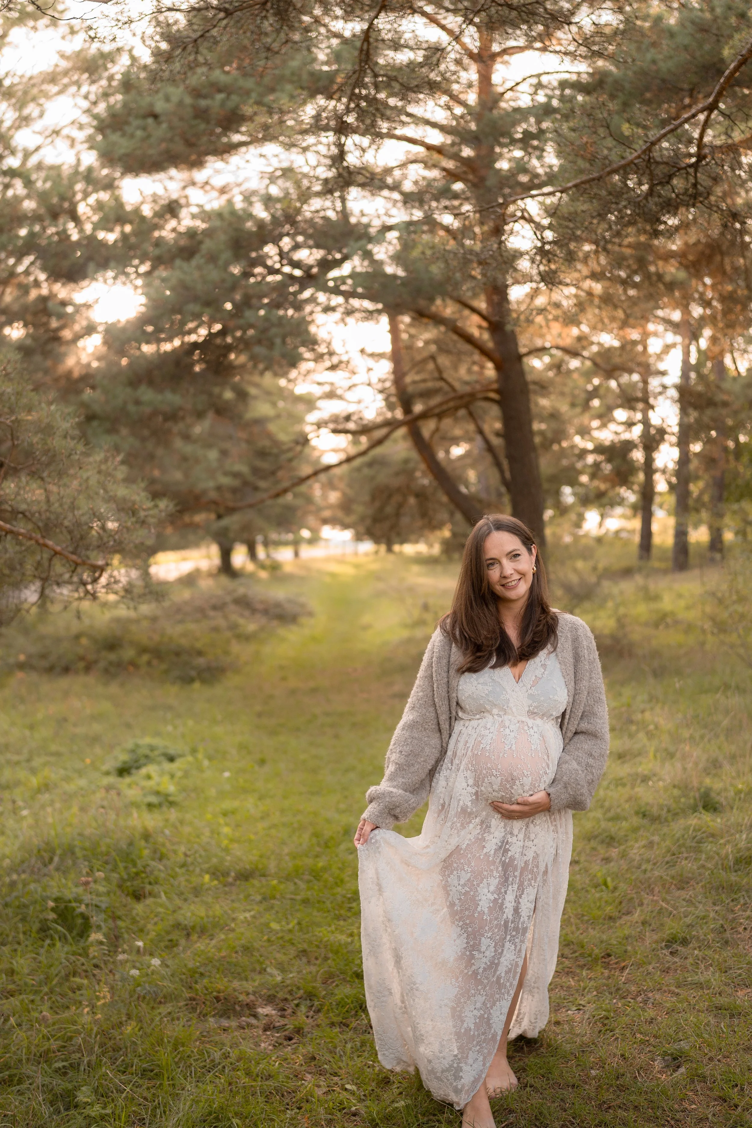 Schwangere Frau in einem weißen, durchsichtigen Kleid und grauem Pullover, die barfuß auf einem Wiesenweg im Sonnenuntergang in einer bewaldeten Gegend steht, lächelnd und die Hand auf ihrem Bauch.