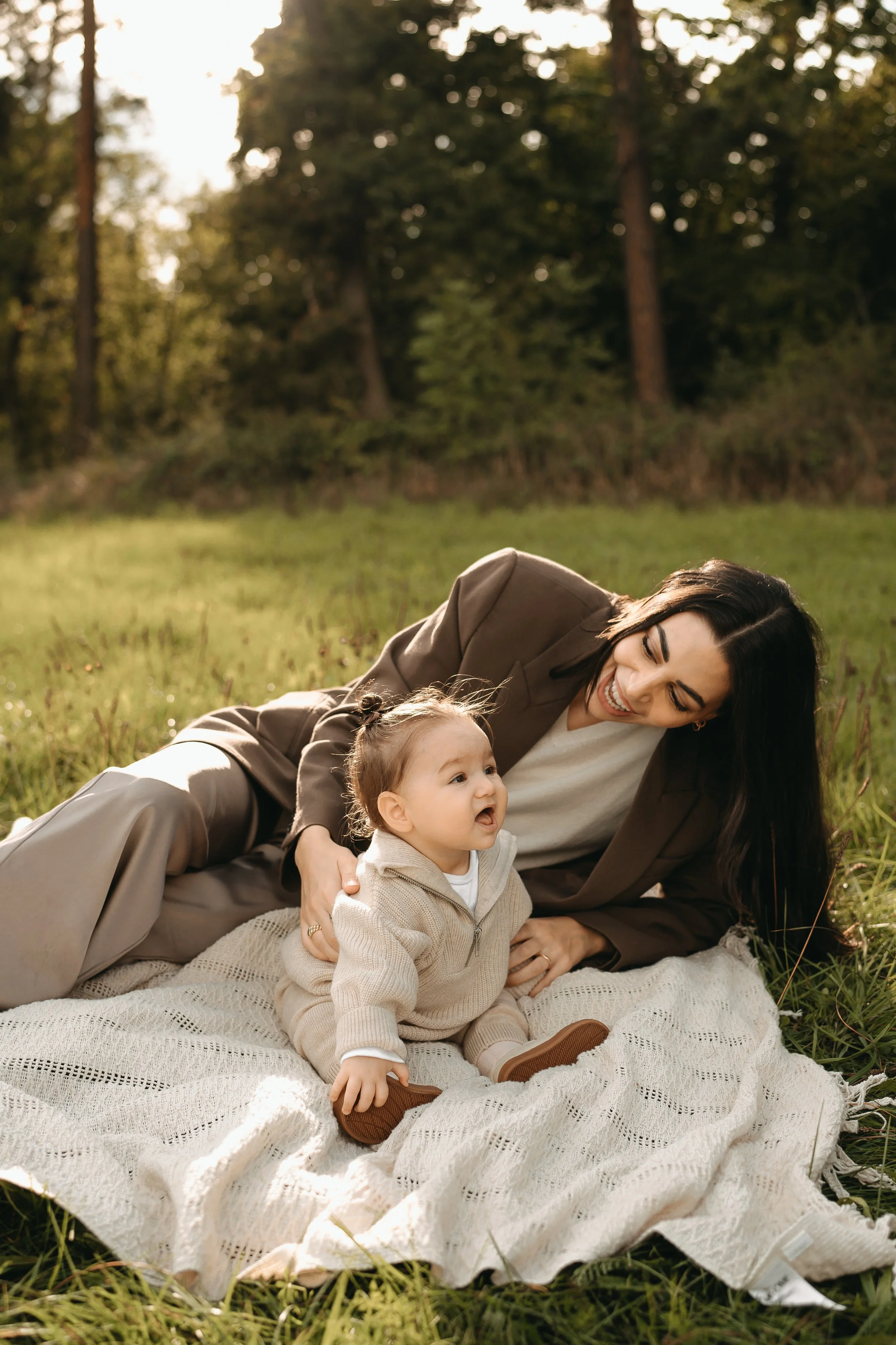 Eine Frau und ein kleines Kind, die auf einem Decken im Freien auf einer Wiese sitzen, lächeln und haben Spaß, umgeben von Bäumen bei Sonnenuntergang.