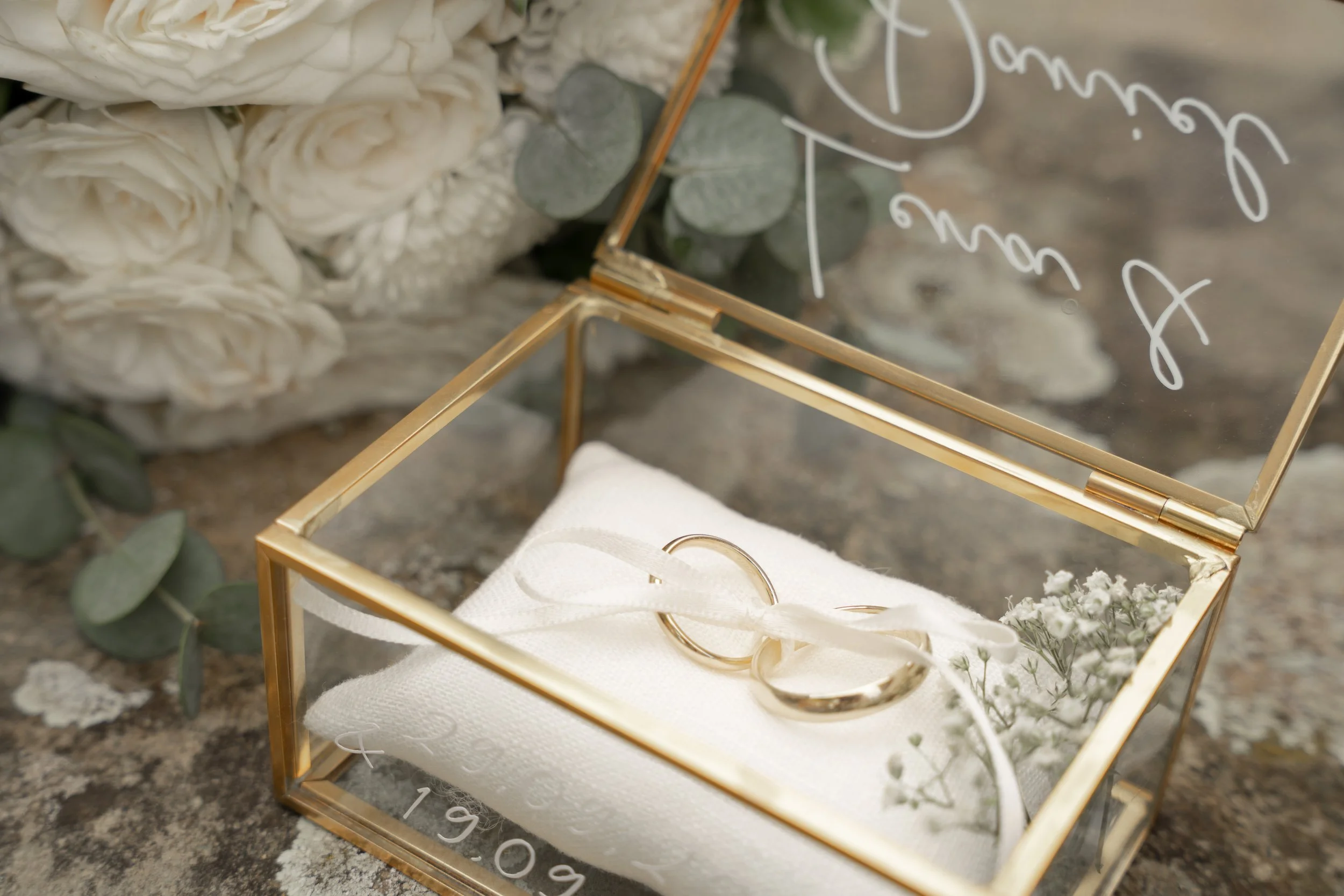 Goldene Ringbox mit zwei Eheringen und Blumen vor einer weißen Rosenbouquet.