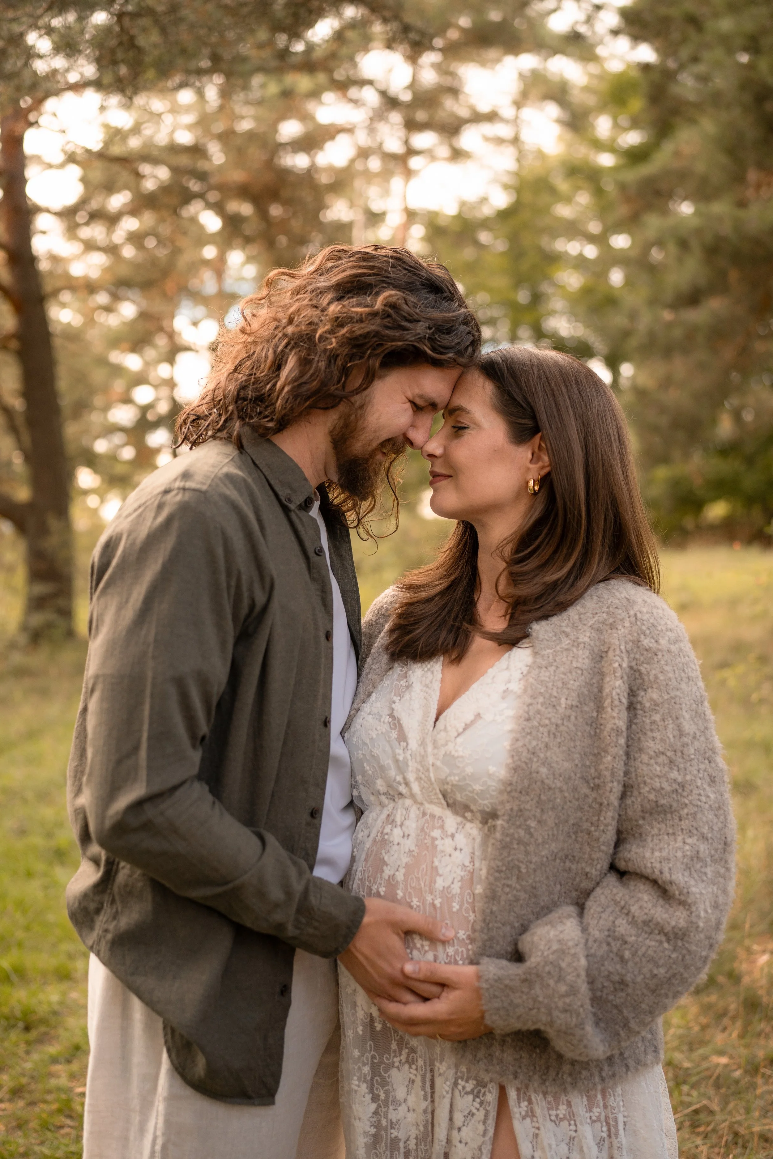 Ein Paar umarmt sich liebevoll in einem Park, sie ist schwanger, beide lächeln sich an, bei Sonnenuntergang, im Hintergrund Bäume.