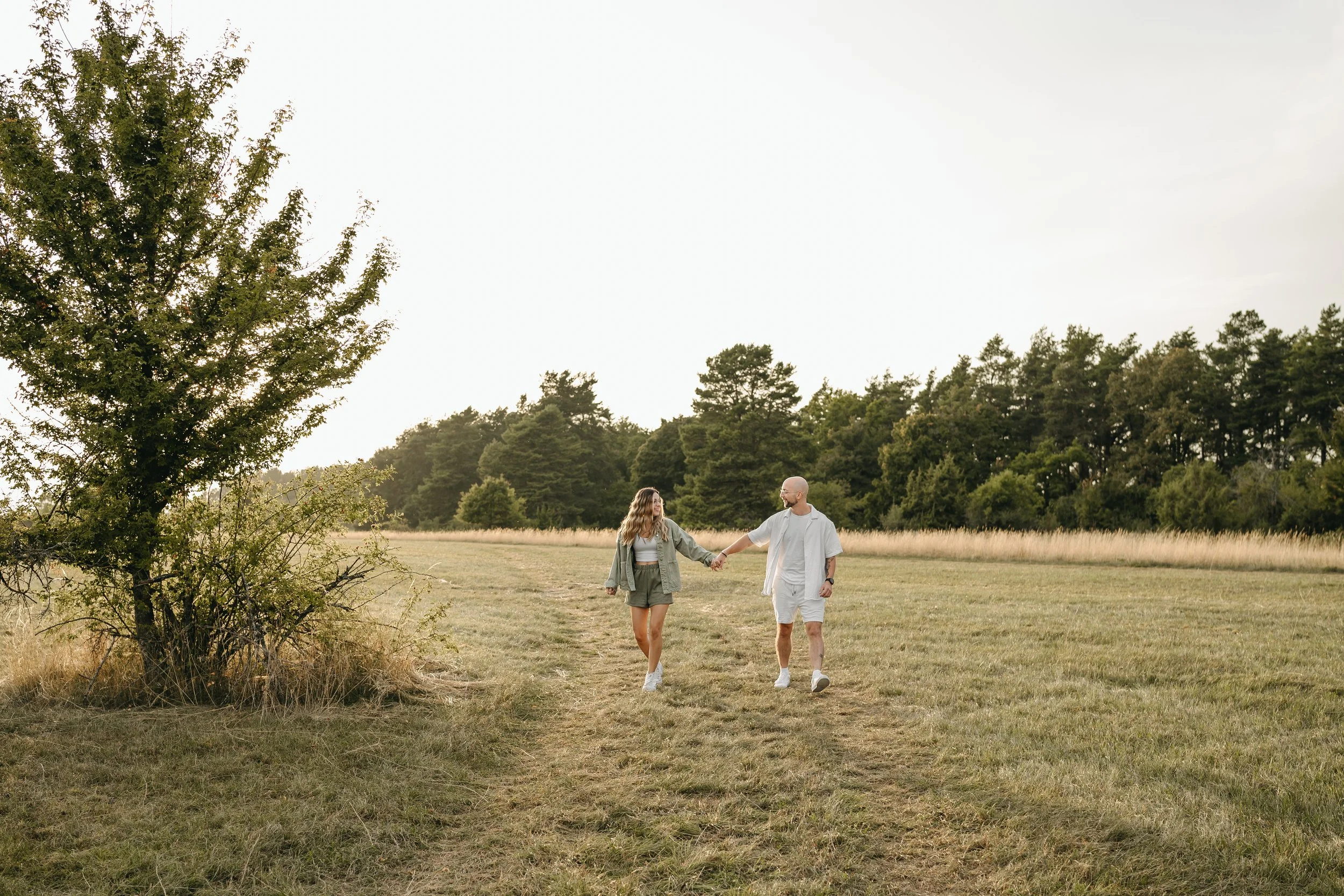 Ein Paar spaziert Hand in Hand durch eine offene Wiese, umgeben von Bäumen bei Sonnenuntergang.