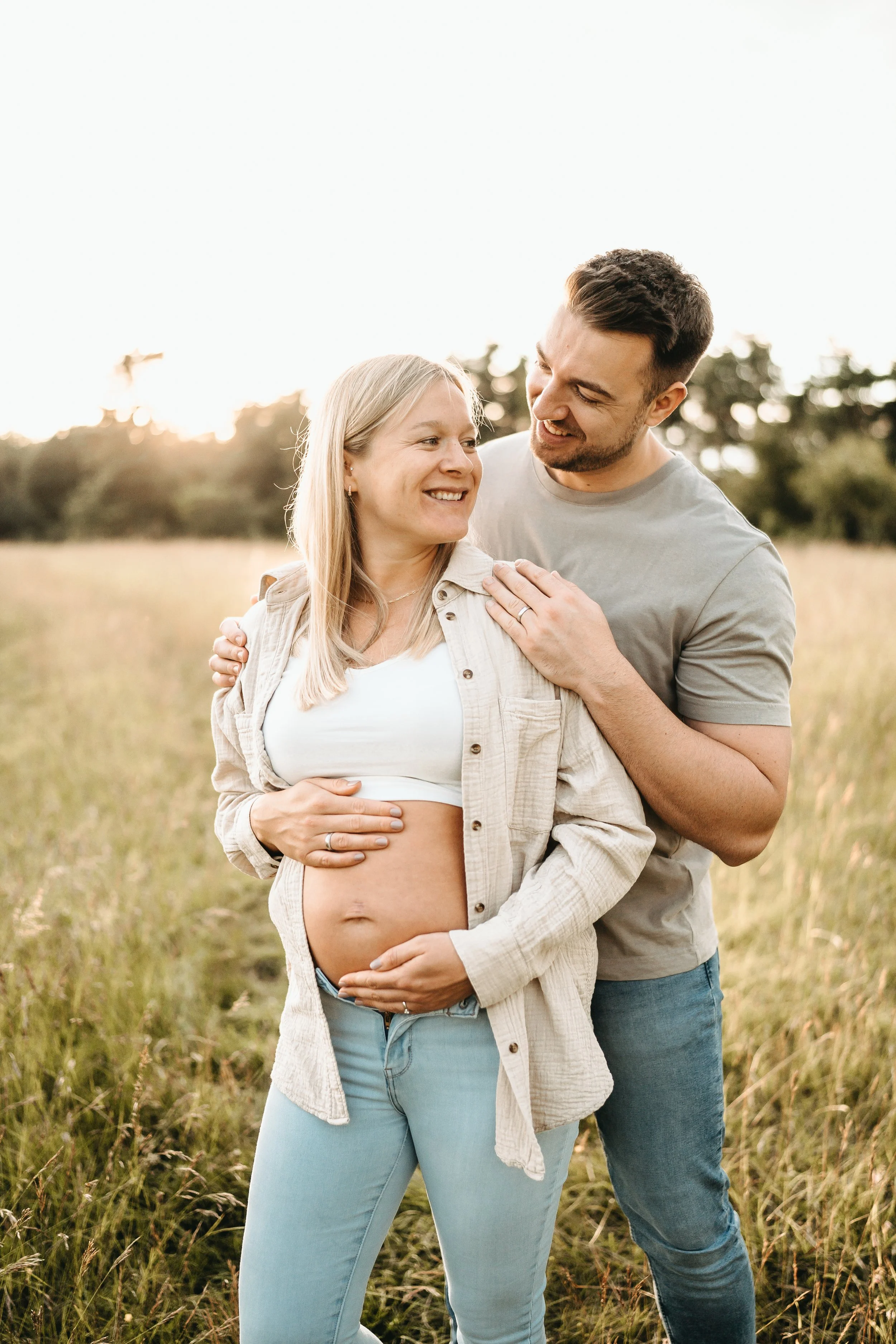 Ein Paar steht in einem Feld bei Sonnenuntergang, die schwangere Frau lächelt, der Mann legt die Hand auf ihre Schulter.