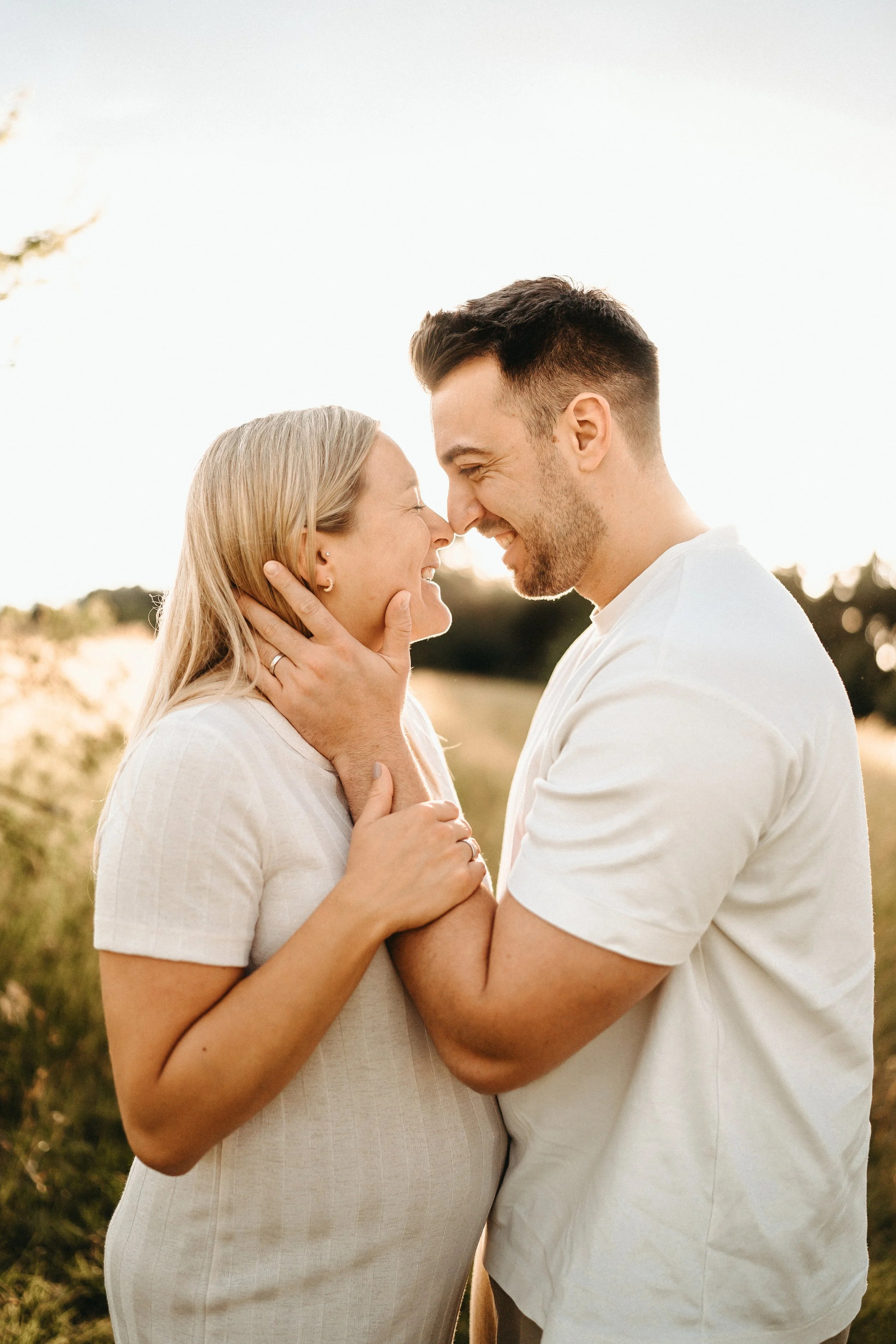 Ein glückliches Paar steht in einer Naturkulisse bei Sonnenuntergang, berühren sich mit der Stirn, lachen und schauen sich liebevoll an.