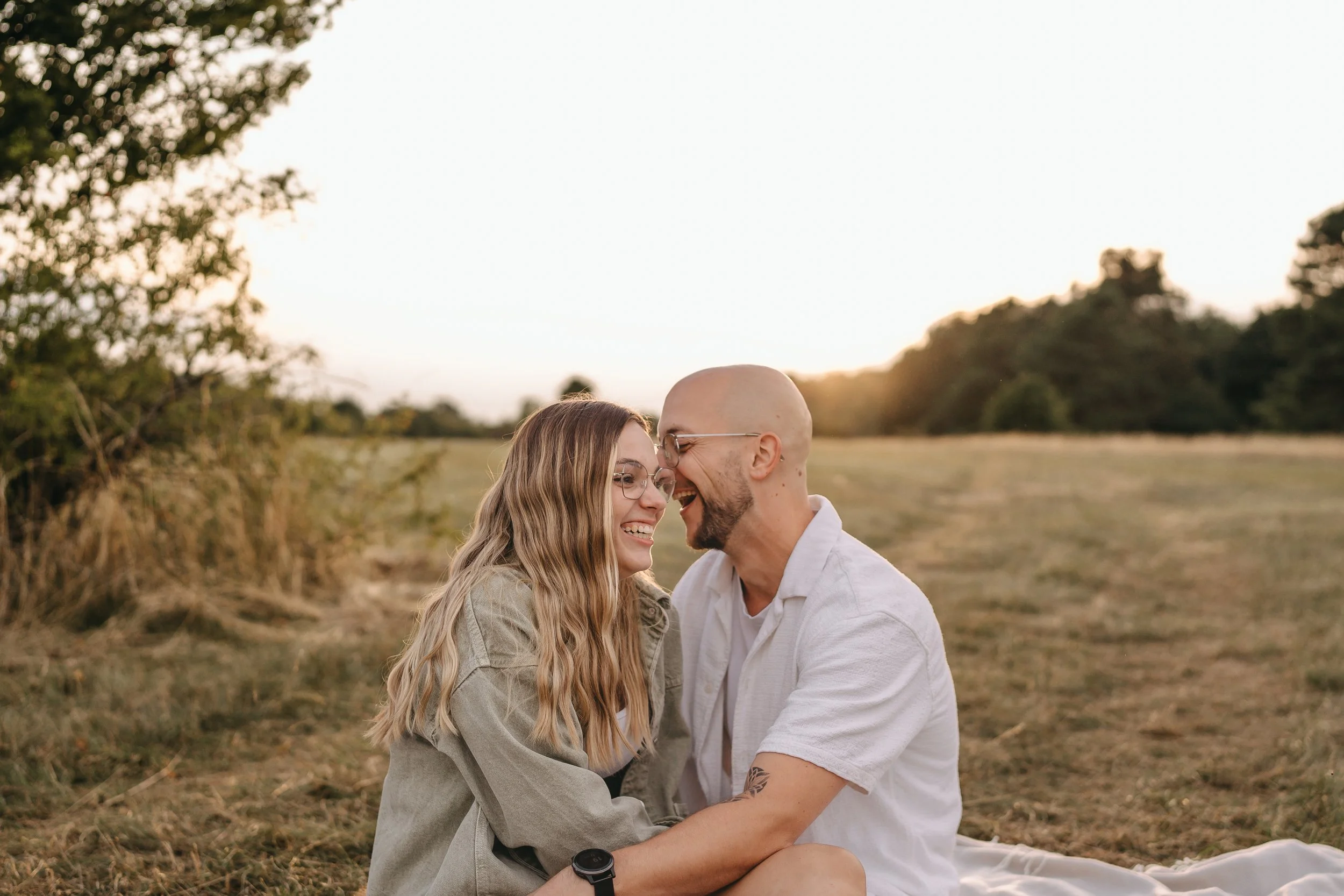 Ein lachendes Paar sitzt im Freien in einer Wiese bei Sonnenuntergang, umgeben von Bäumen.