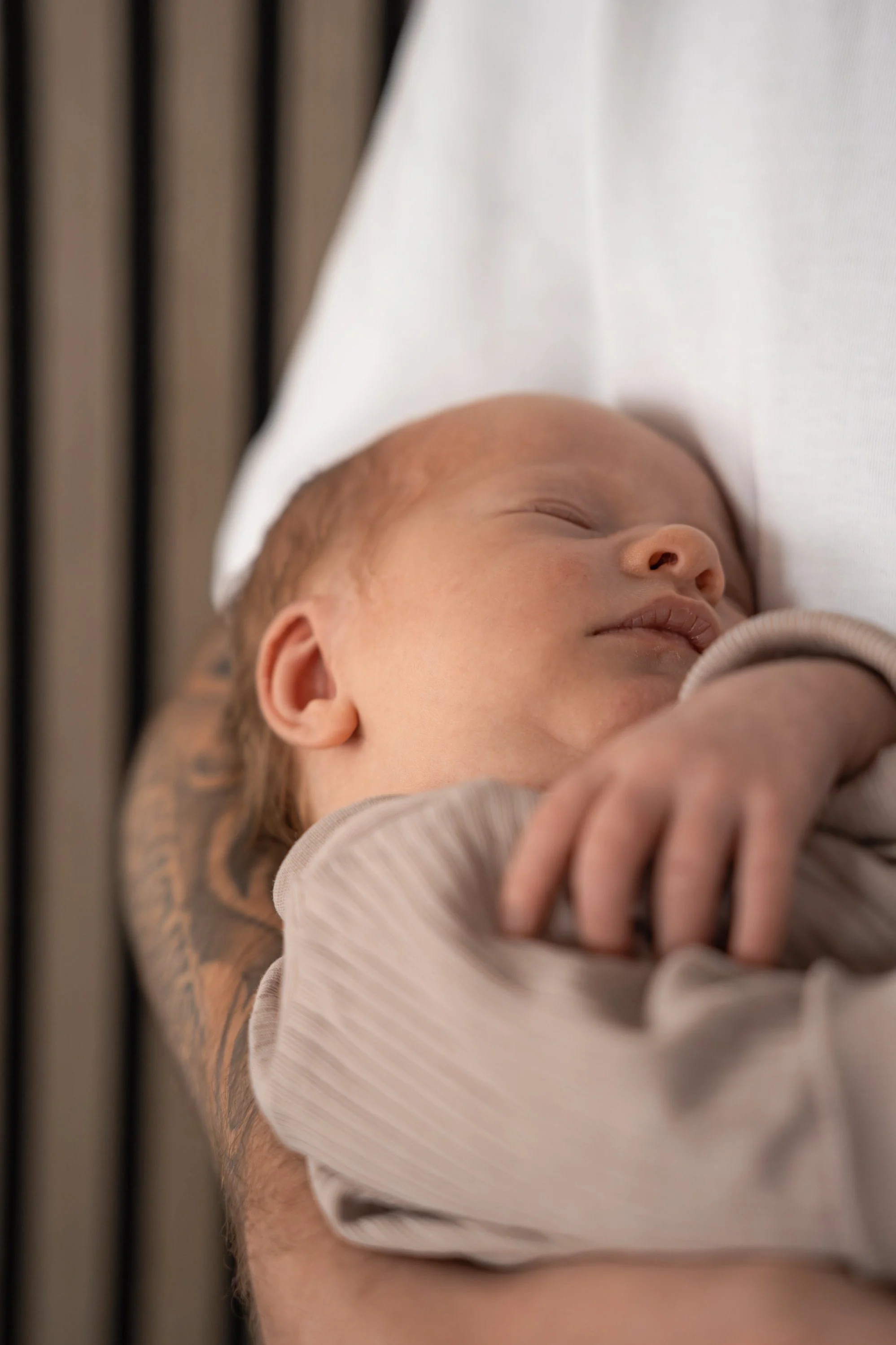 Ein schlafender Säugling wird von einer Person mit tätowiertem Arm gehalten, während sie auf einem Bett liegt.
