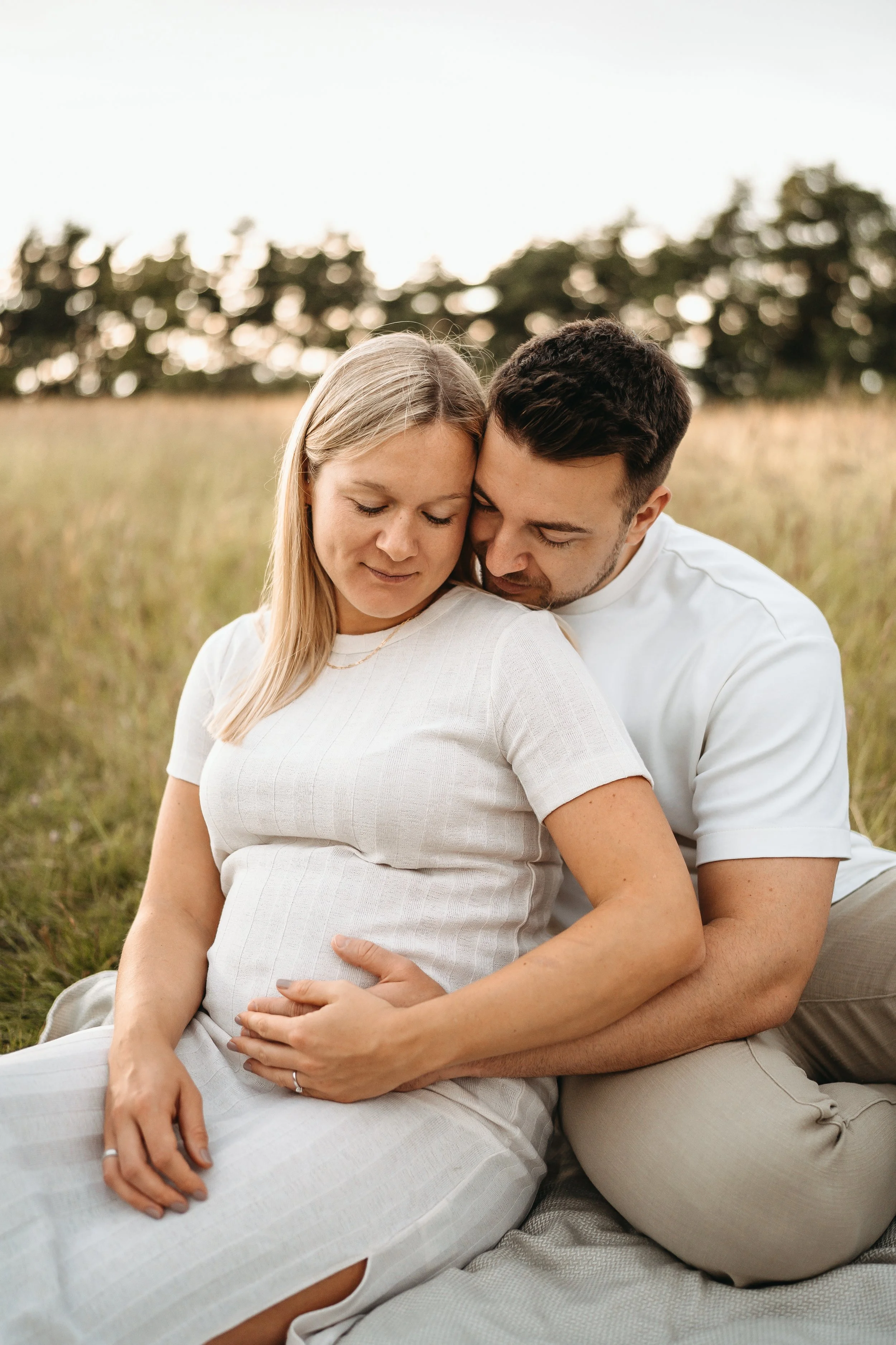 Ein schwangere Frau und ein Mann sitzen im Gras, umarmen sich und schauen liebevoll auf den Bauch der Frau in einem offenen Feld bei Sonnenuntergang.