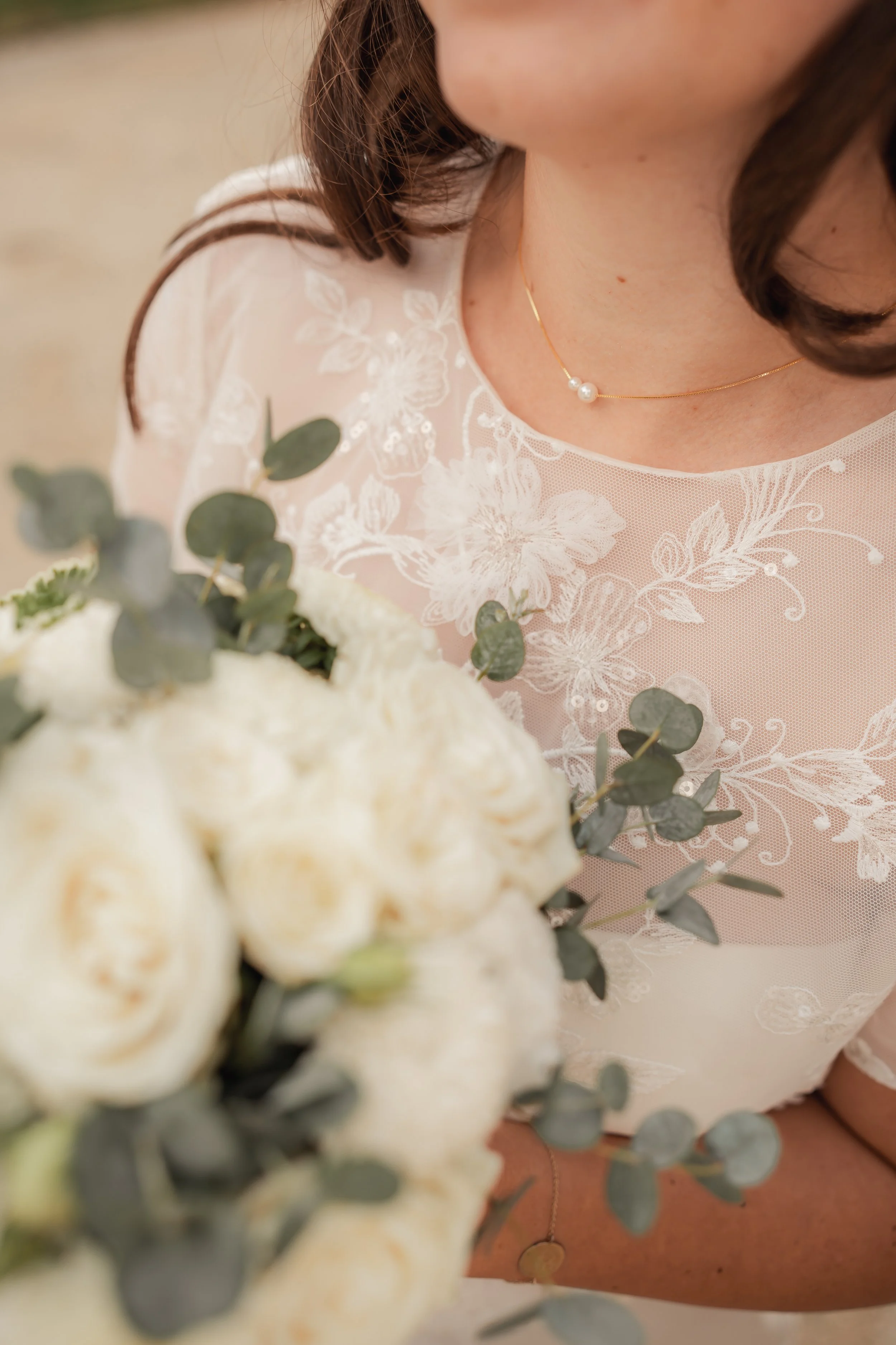 Nahaufnahme einer Braut mit Perlenkette, Trägerin eines Spitzenkleids mit Blumenmuster, hält einen Blumenstrauß aus weißen Rosen und Eukalyptus.