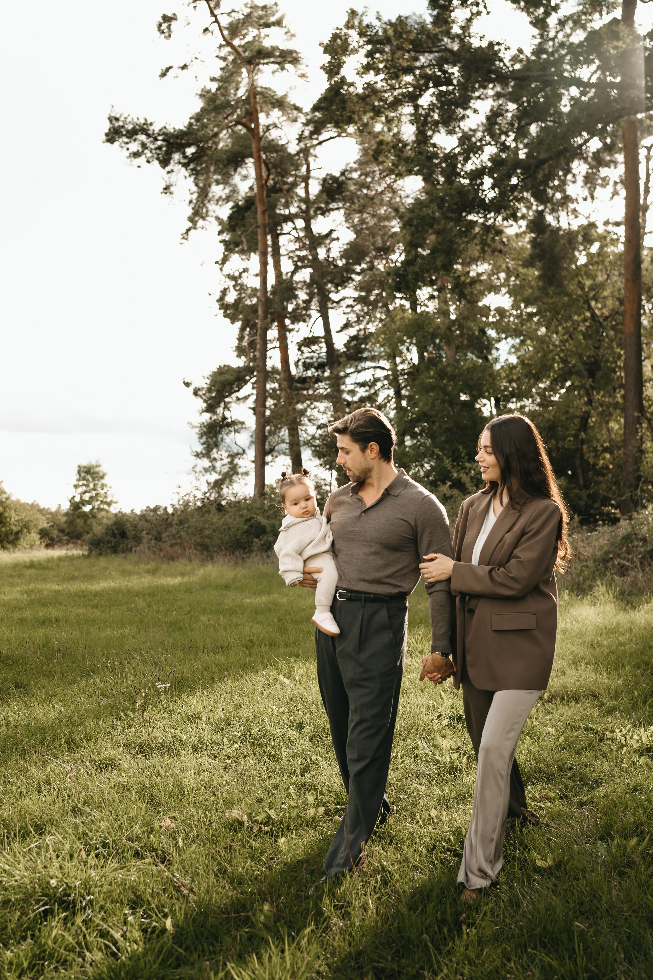 Ein Mann hält ein kleines Mädchen, während eine Frau sie an der Hand hält, im Freien auf einer Wiese mit Bäumen im Hintergrund bei Sonnenuntergang.