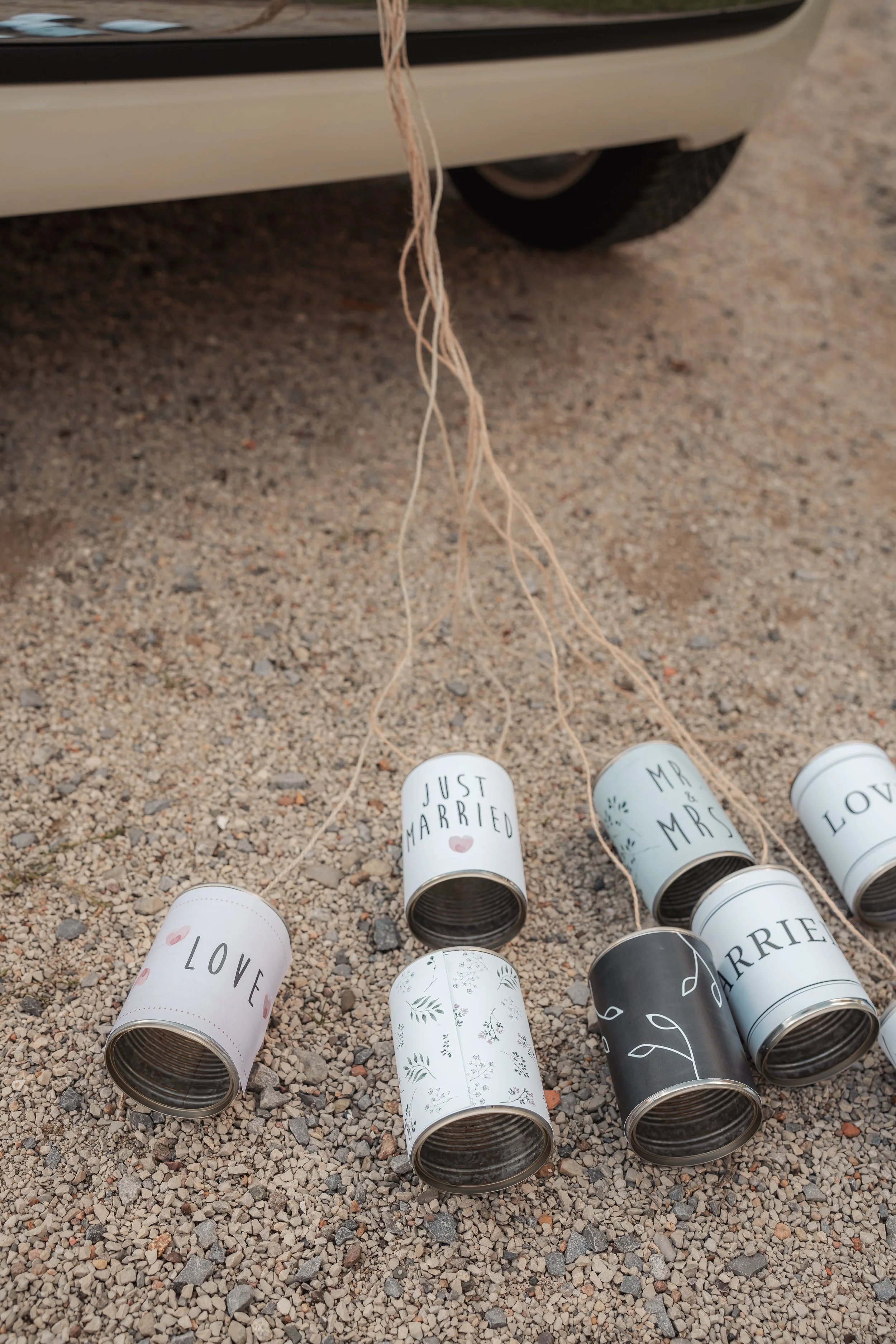 Leere Dosen mit Hochzeits- und Liebe-Botschaften auf Kiesboden, mit Auto im Hintergrund.