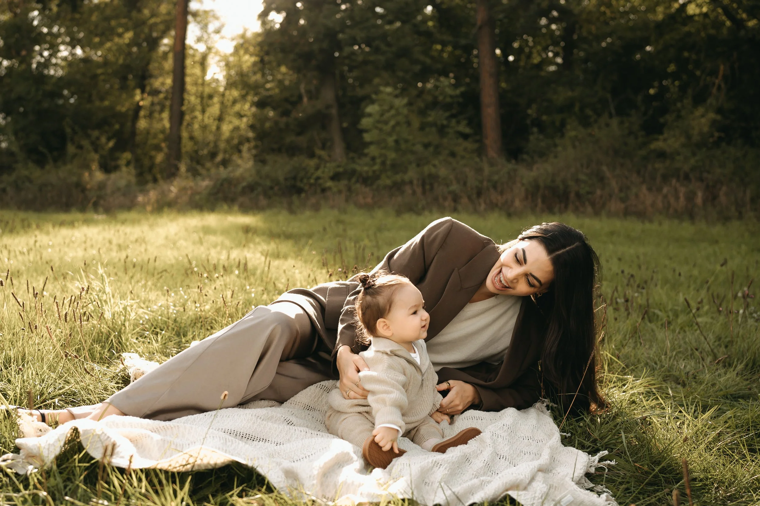 Eine Frau liegt mit einem Baby auf einem weißen Tuch im Gras beim Picknick im Wald, beide lachen und genießen die sonnige Umgebung.