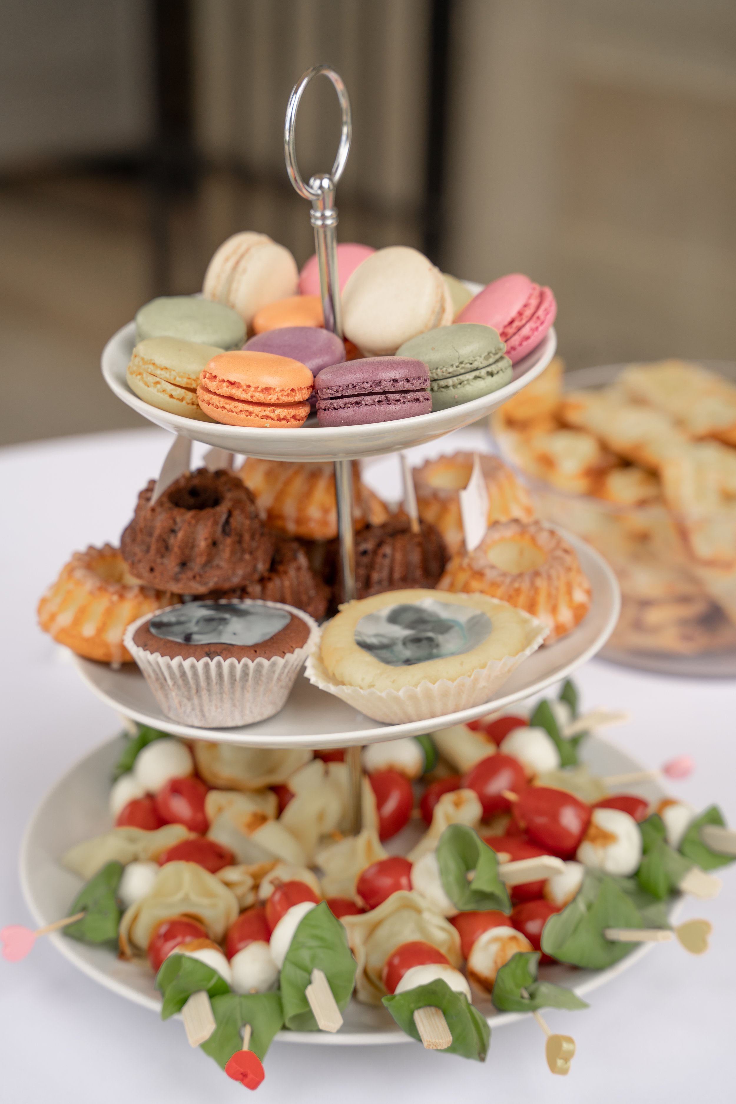 Verschiedene Desserts und Kekse auf einem mehrstöckigen Desserttablett, inklusive Macarons, Cupcakes, Cookies und Vorspeisen mit Tomaten und Käse.
