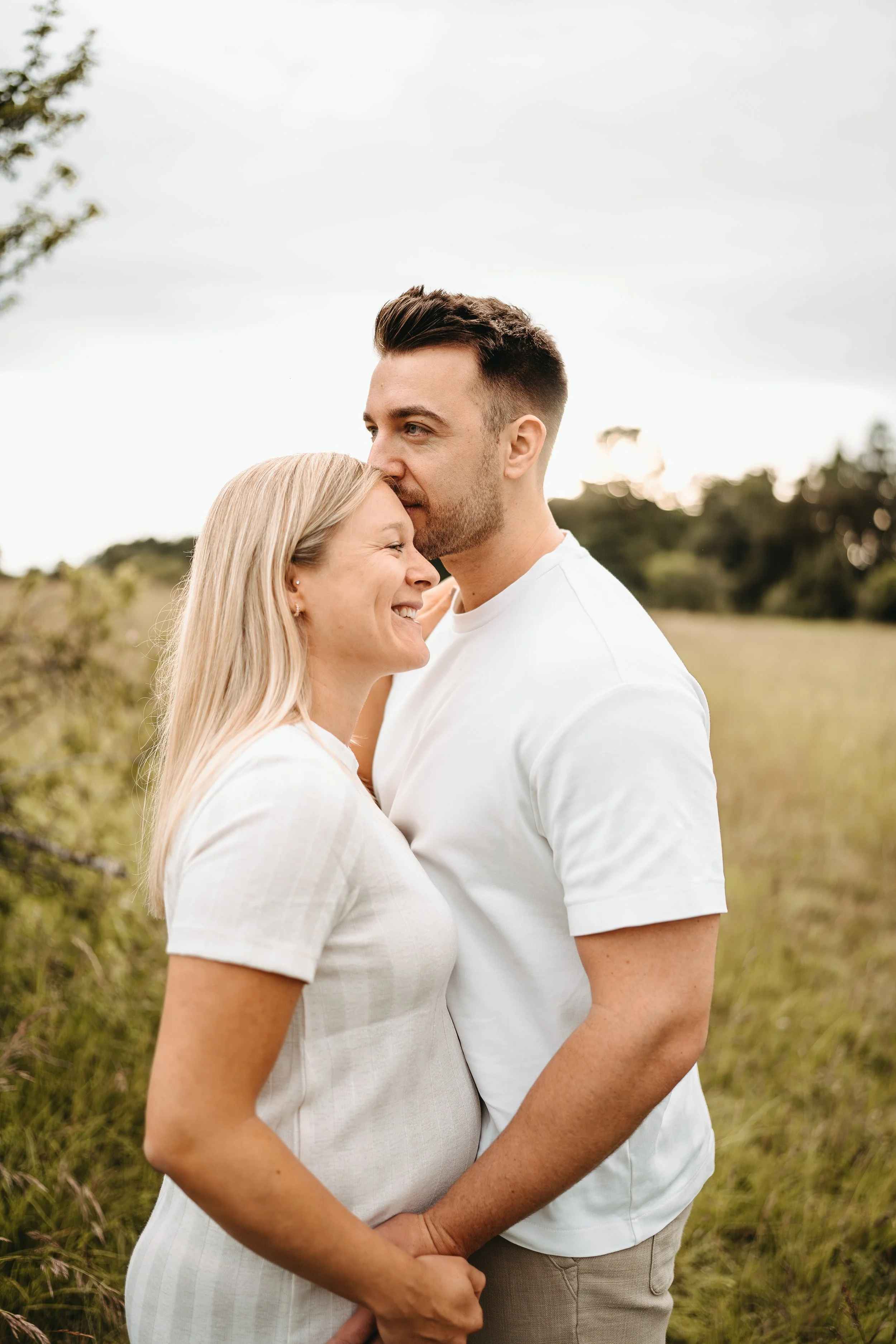 Ein Paar steht eng vor einem grünen Feld, lächelt und umarmt sich.