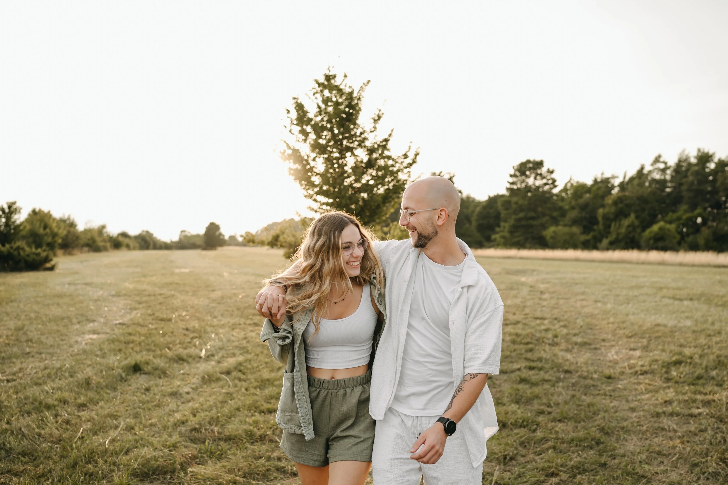 Ein lachendes Paar geht umarmend durch eine grüne Wiese bei Sonnenuntergang, umgeben von Bäumen.