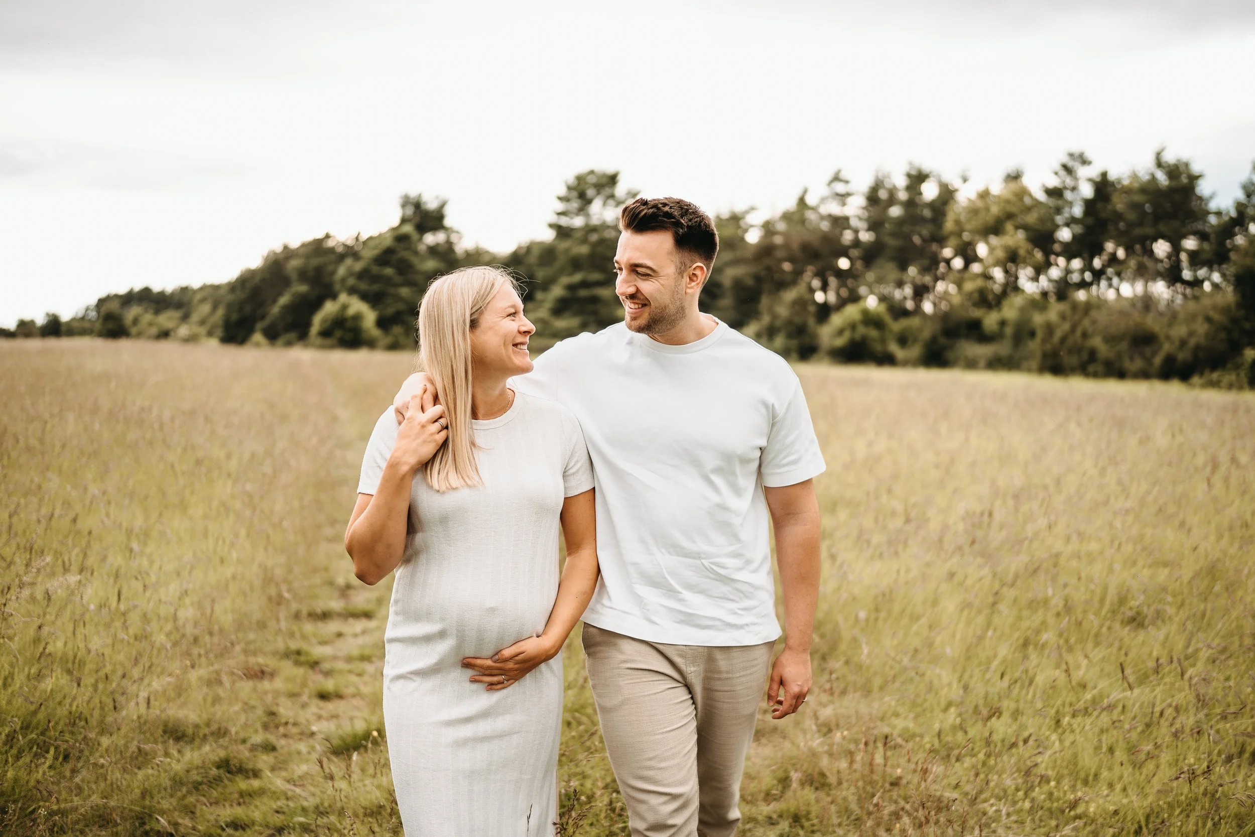 Ein lachendes Paar in einem Feld bei bewölktem Himmel, die Frau legt eine Hand auf den Bauch, die Männer schaut sie an.