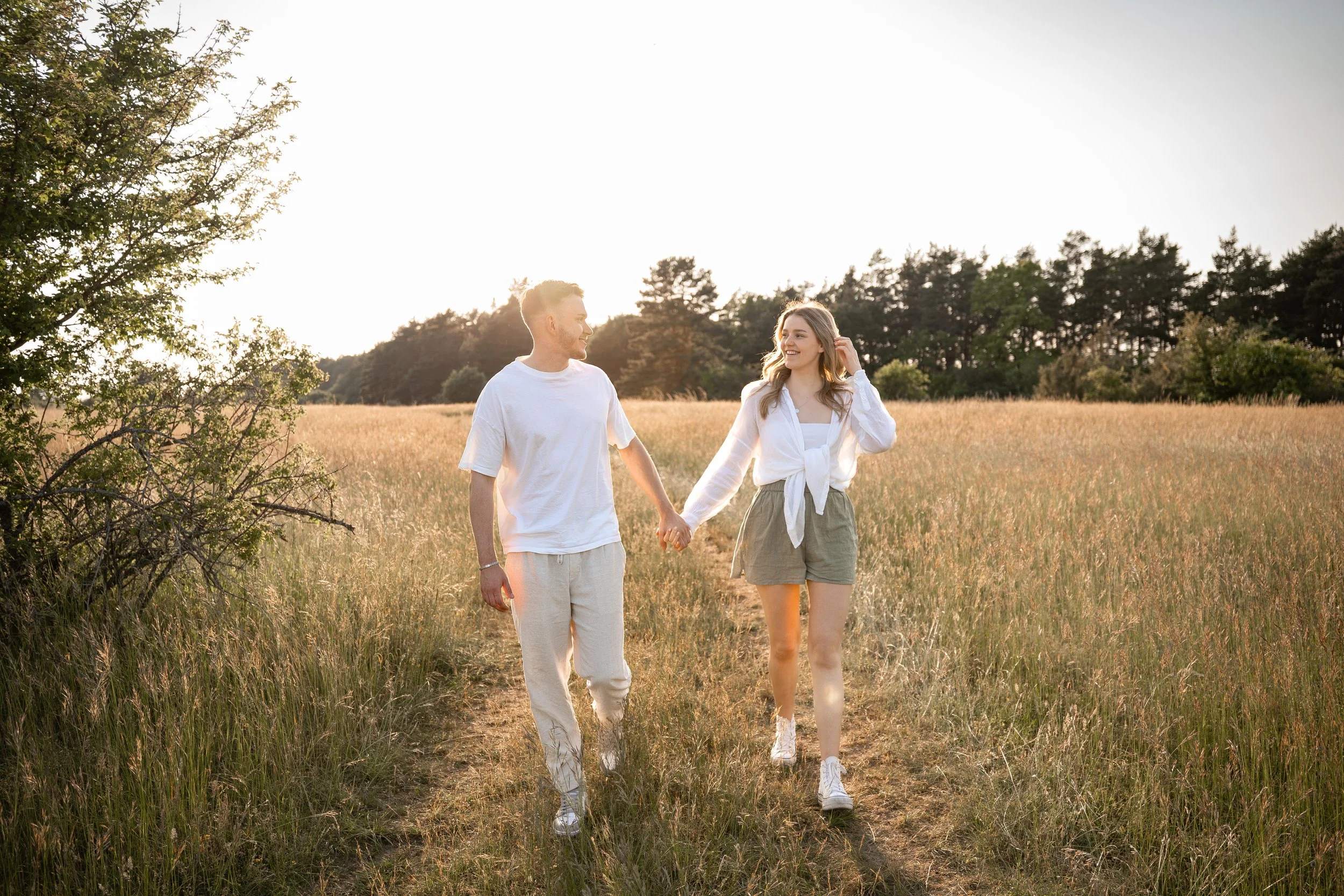 Ein junges Paar spaziert Hand in Hand durch eine sonnige Wiesenlandschaft bei Sonnenuntergang.
