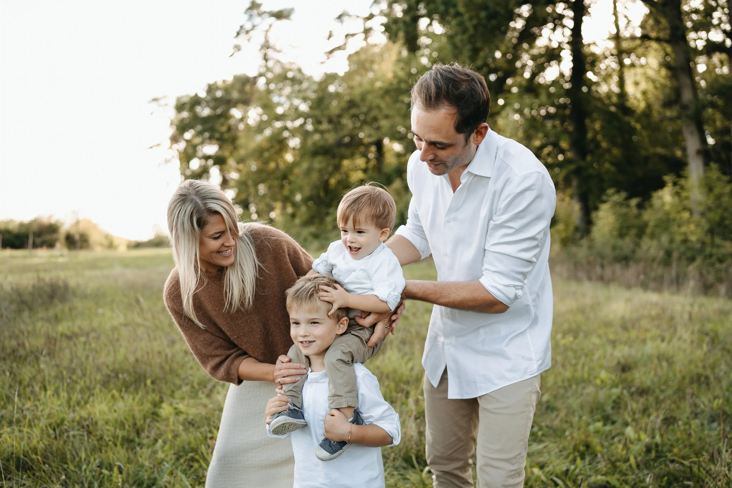 Familie mit zwei Kindern spielt lachend im Grünen, während die Eltern die Kinder liebevoll halten. Es ist ein sonniger Tag im Freien mit Bäumen im Hintergrund.