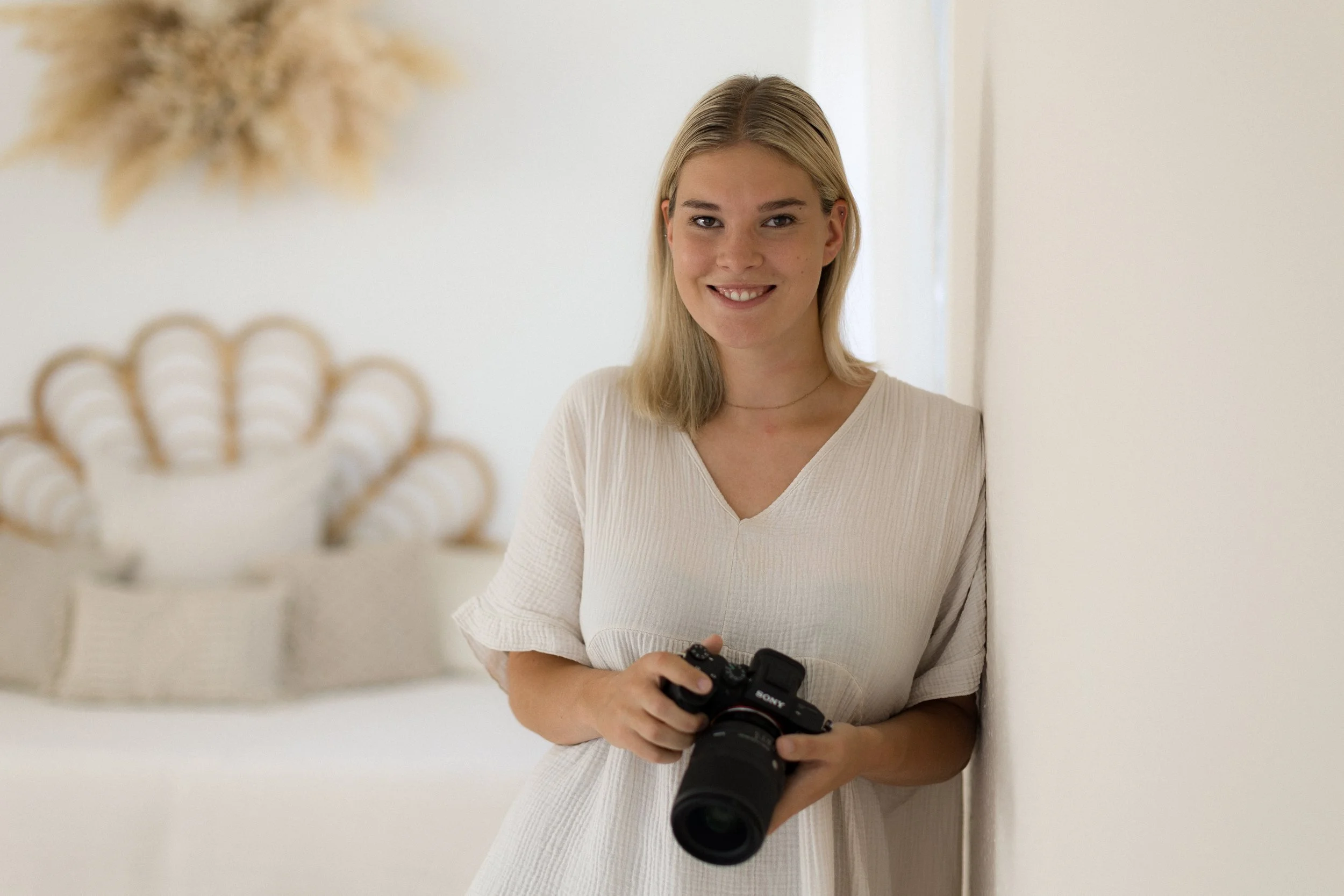 Junge Frau mit Kamera lehnend an weiße Wand im hellen Raum, unscharf dekorative Wandkunst im Hintergrund.