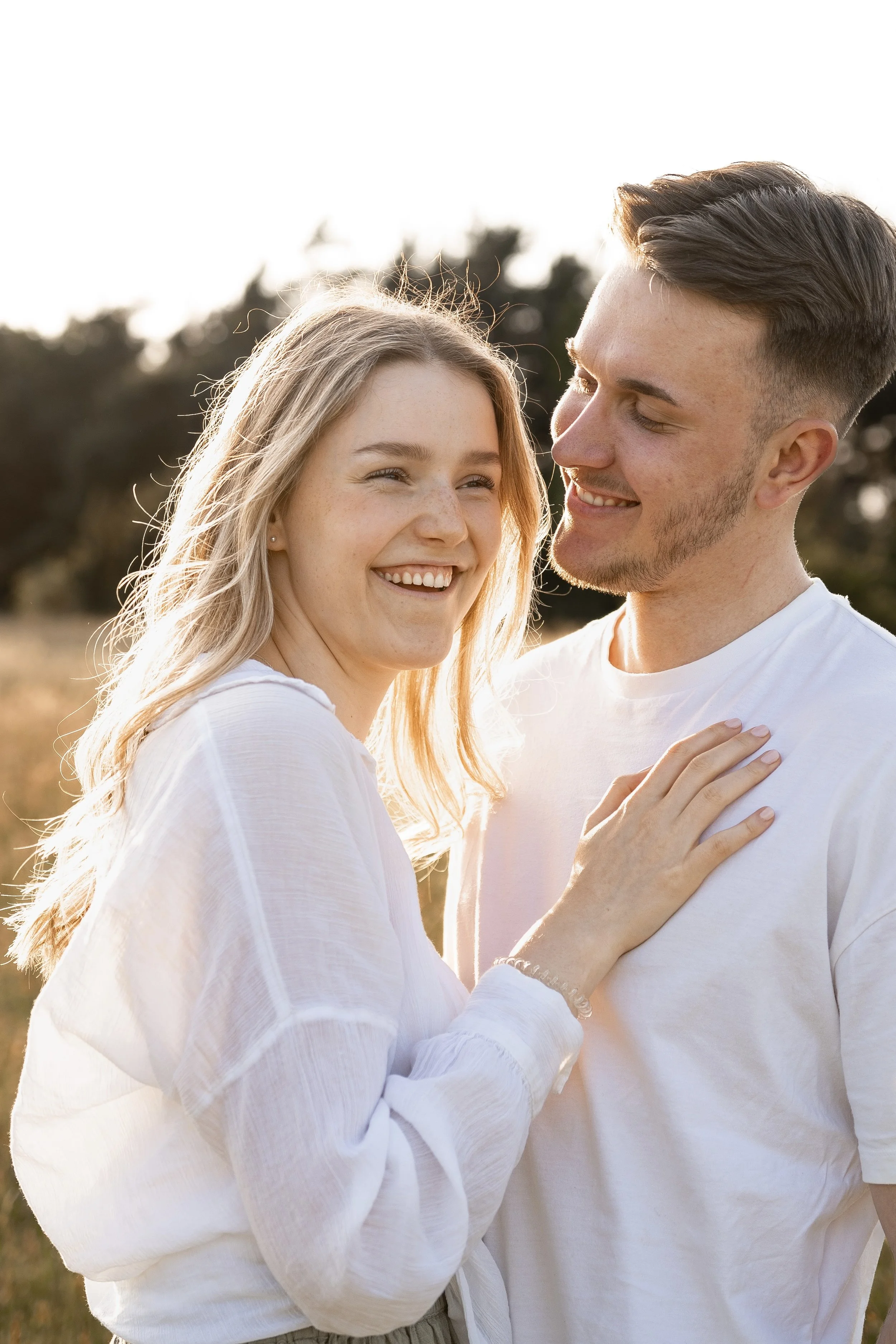 Ein lachendes Paar steht draußen im Sonnenlicht, umgeben von Natur, und scheint glücklich und verliebt.