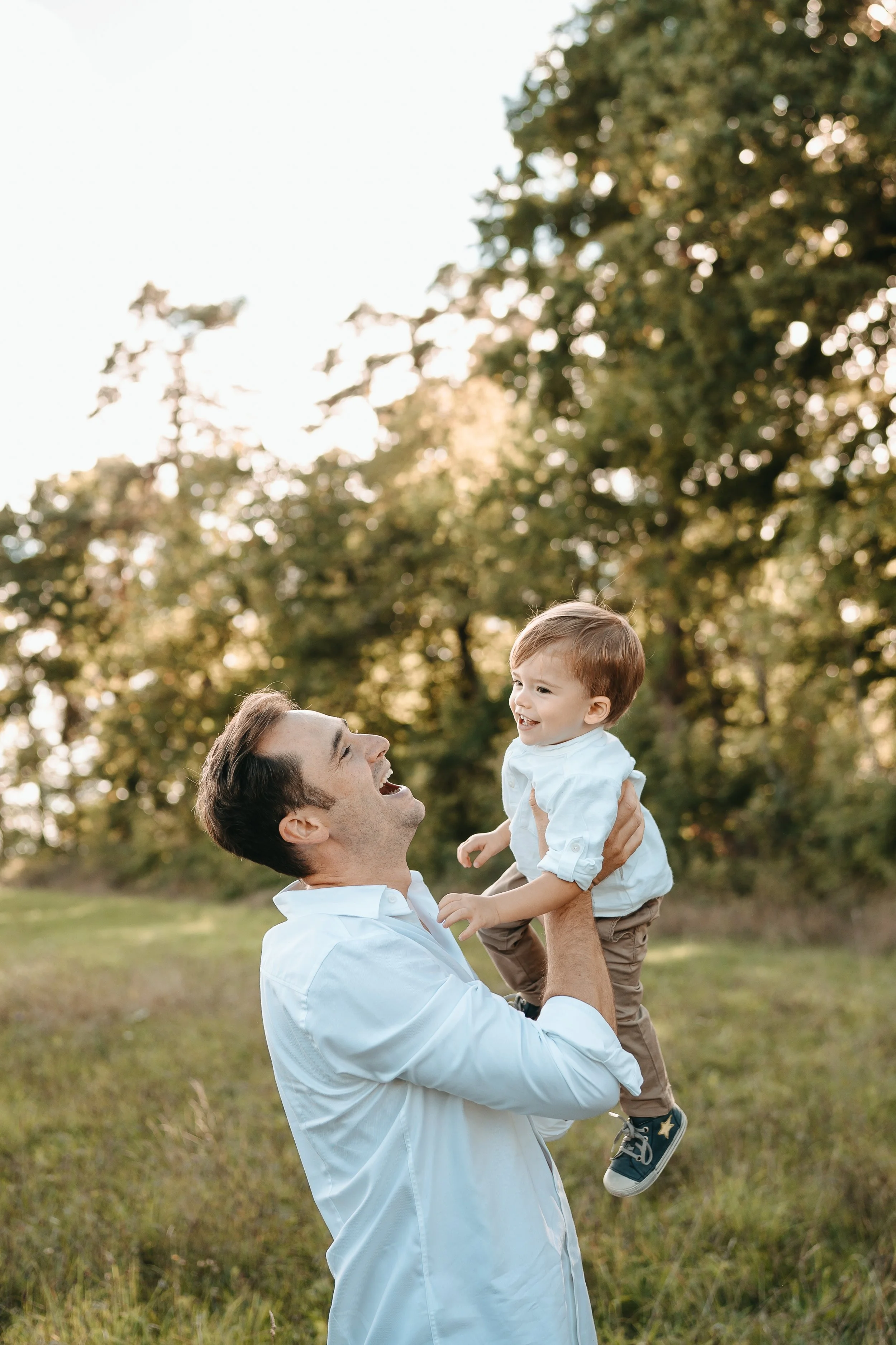 Vater hebt kleinen Jungen in einem Park hoch, beide lachen und genießen die Natur bei Sonnenuntergang.