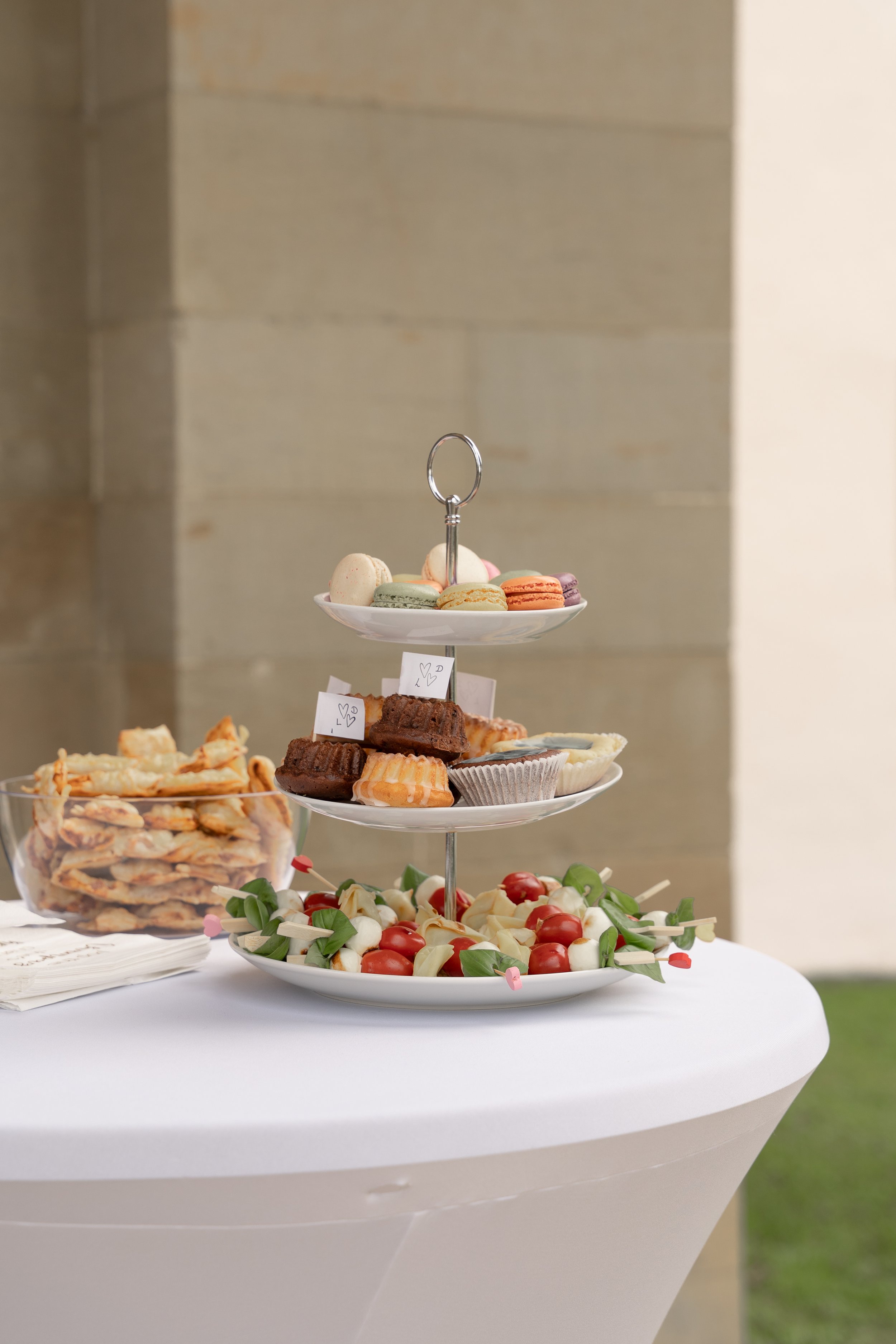 Ein zweistöckiges Buffett mit Macarons, Cupcakes, Sandwiches, Tomaten und Käse auf einem weißen Tischtuch.