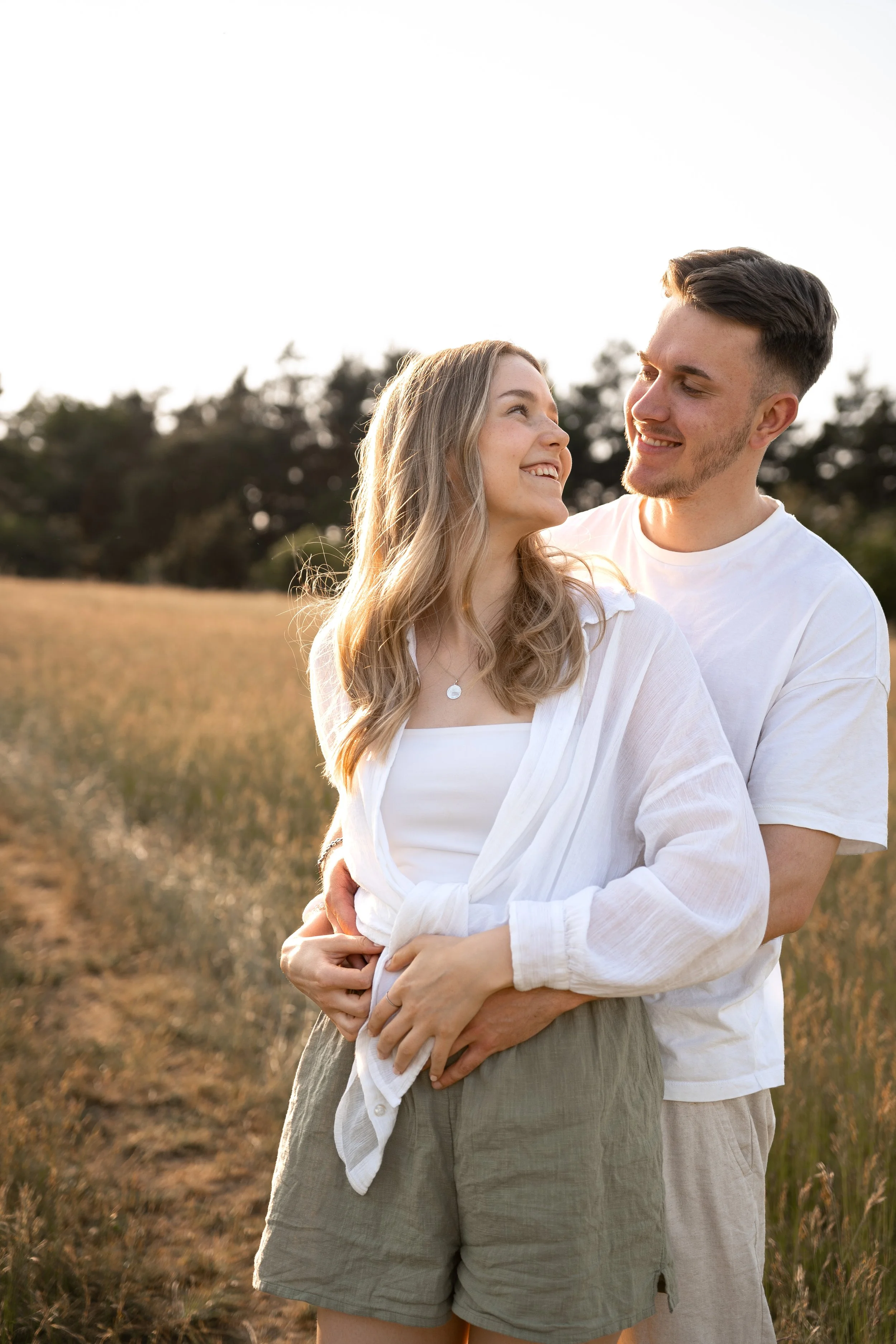 Ein junges Paar umarmt sich in einem Feld bei Sonnenuntergang, beide lächeln sich an.