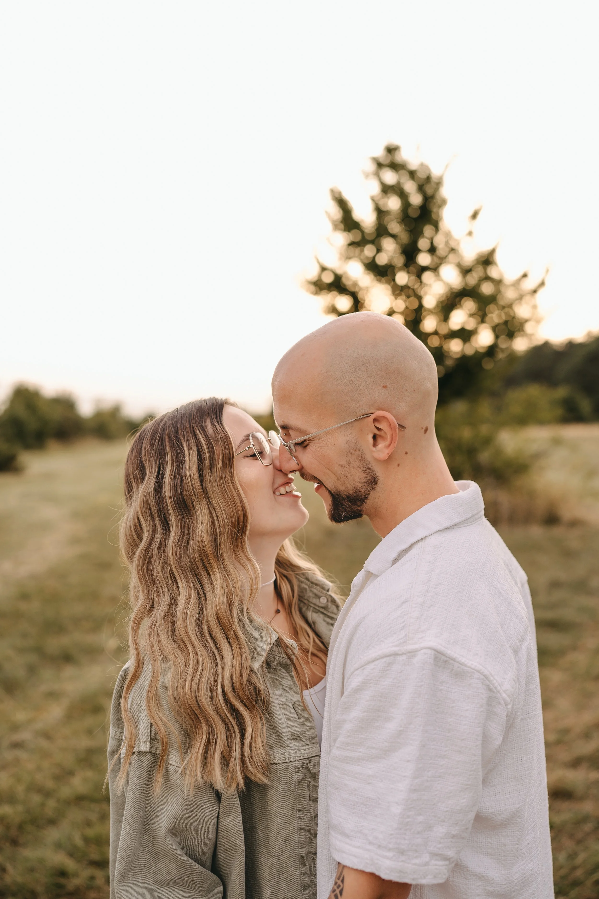 Ein verliebtes Paar steht sich in einer Naturlandschaft mit Bäumen und offenem Himmel gegenüber, lächelnd mit der Stirn aneinander.