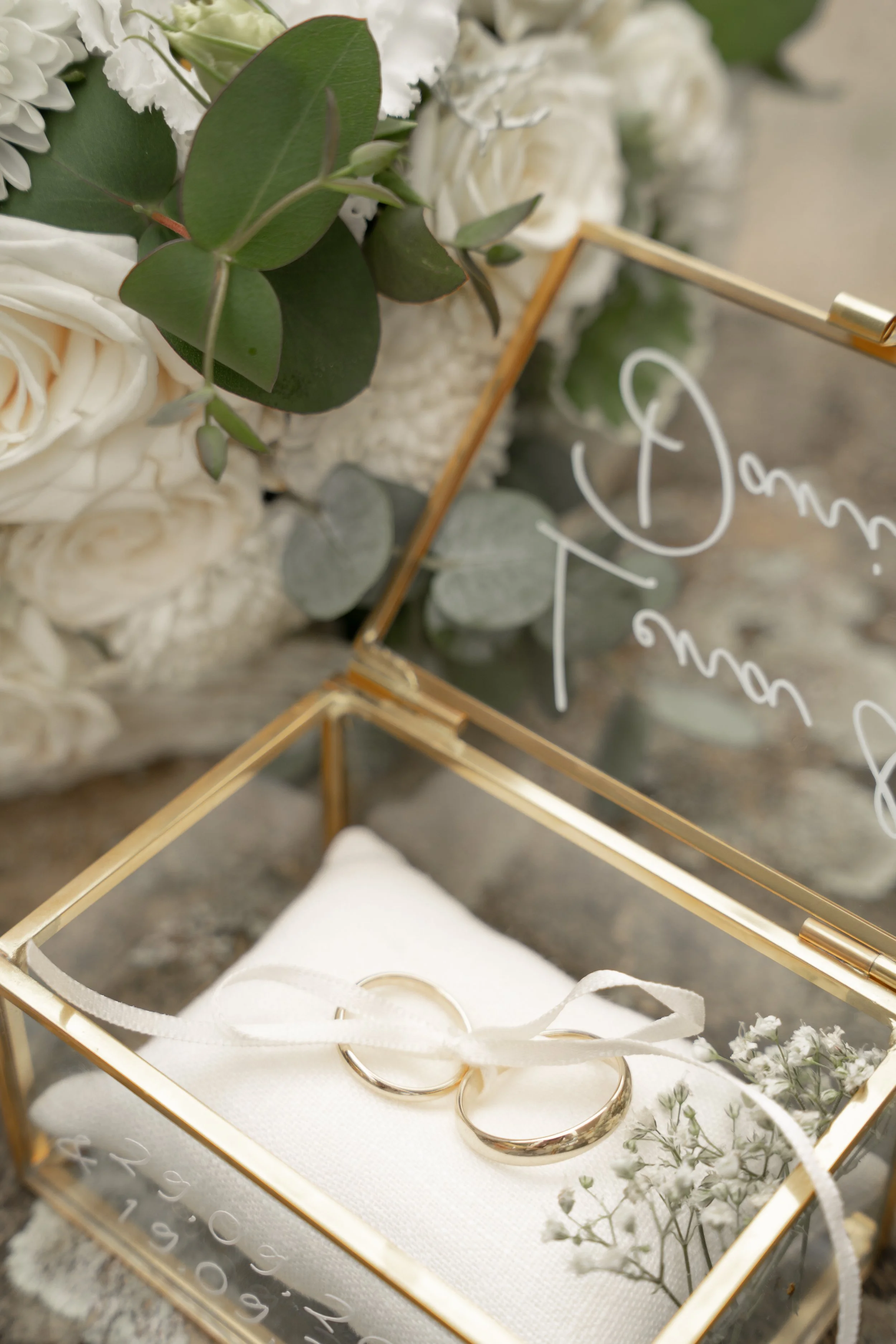 Goldene Schatulle mit Eheringen, umgeben von weißen Blumen und grünem Eukalyptus, mit Hochzeitswünschen als Beschriftung im Hintergrund.