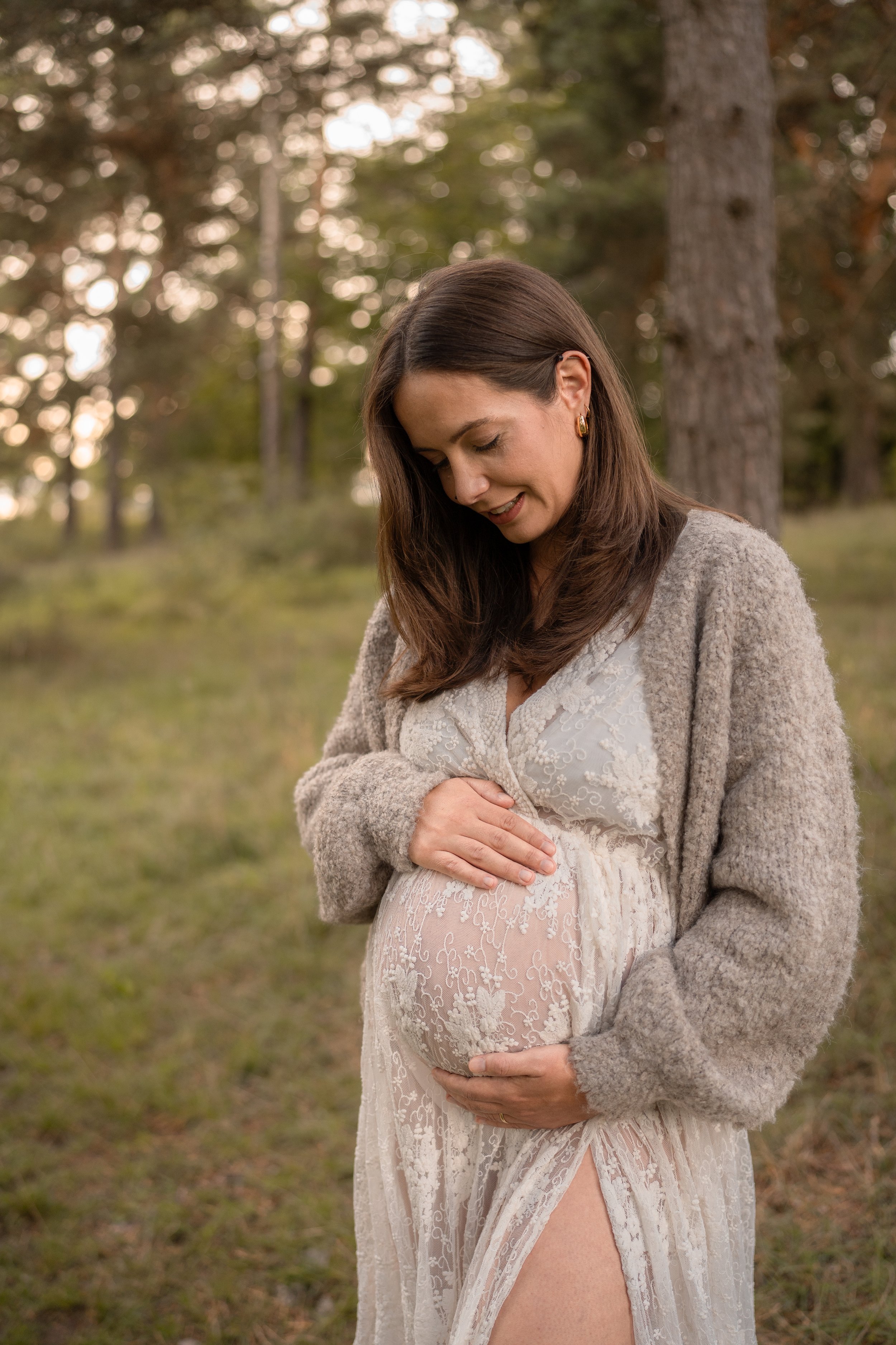 Eine schwangere Frau steht in einem Wald und hält ihre Hand auf ihren Babybauch, während sie nach unten schaut.