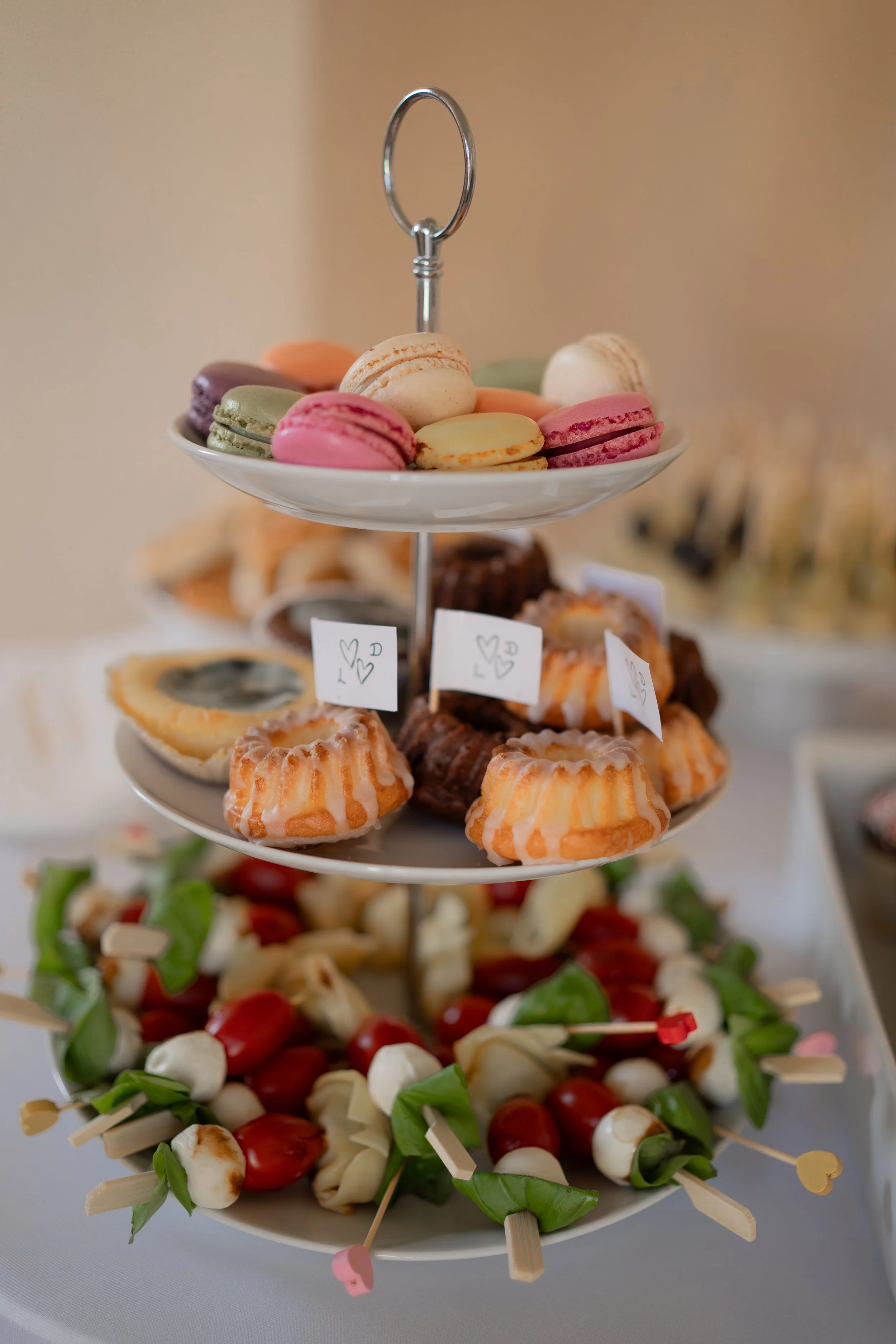 Ein mehrstöckiges Serviertablett mit bunten Macarons oben, kleinen Kuchen in der Mitte und Antipasti-Spießen unten.