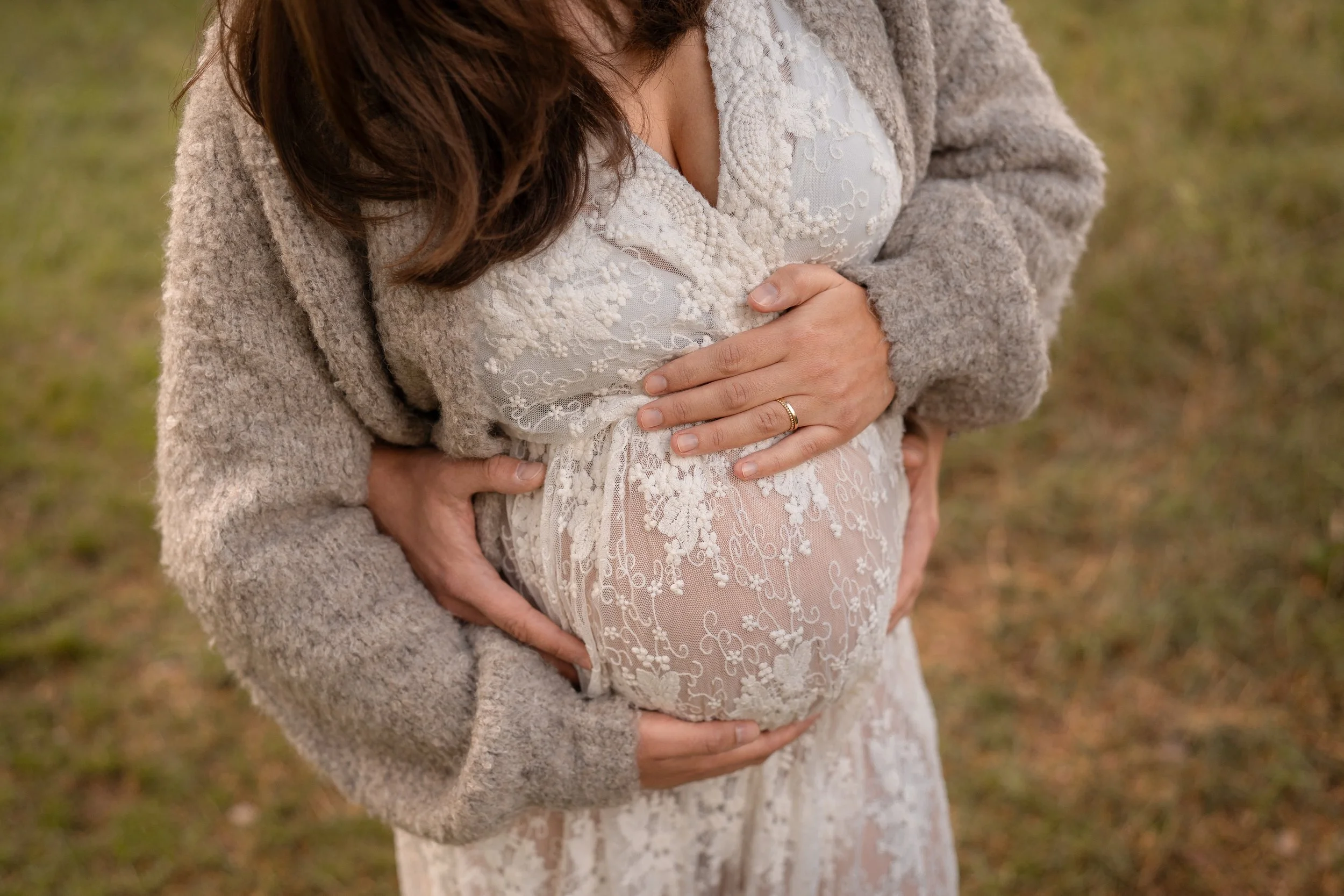 Eine schwangere Frau in einem weißen Spitzenkleid, umarmt ihren Bauch, während sie draußen im Grünen steht, umgeben von warmem Licht.