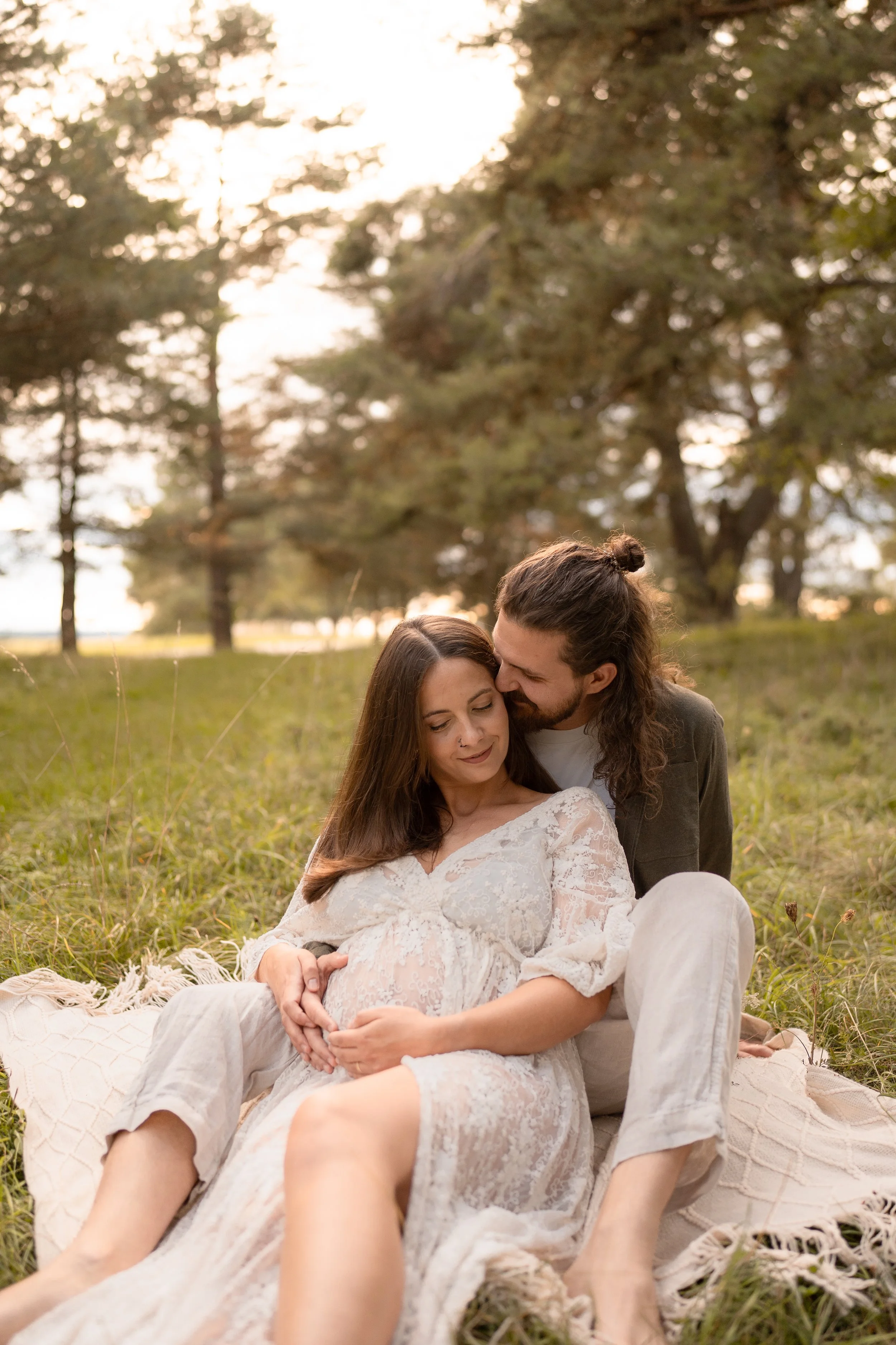 Ein Paar sitzt auf einer Decke im Grünen, die schwangere Frau trägt ein weißes, durchscheinendes Kleid, der Mann küsst sie, umgeben von Bäumen bei Sonnenuntergang.