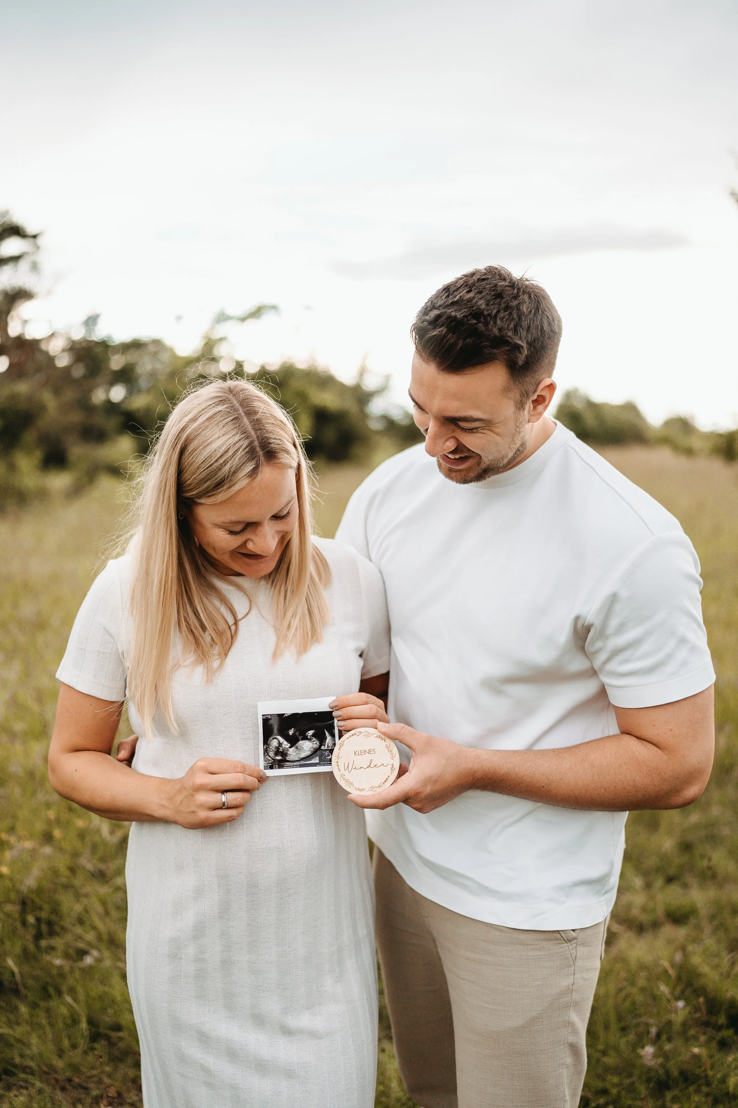 Ein Paar hält einen Ultraschallbild und einen kleinen Holzanhänger mit der Aufschrift 'Kleines Wunder' in der Hand, während es lächelnd auf einem Feld steht.