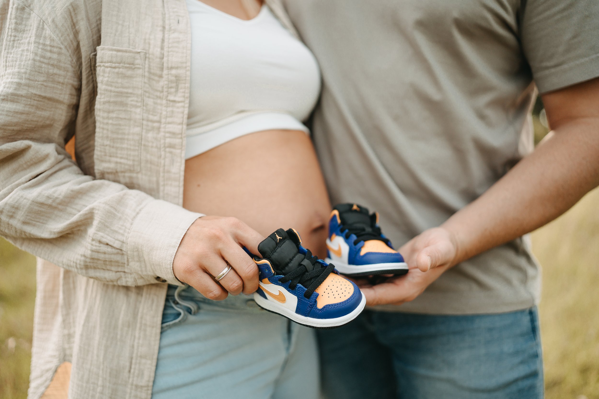 Schwangere Frau hält kleine Sneakers, während ein Mann sie an ihren Beinen anfasst, im Freien.