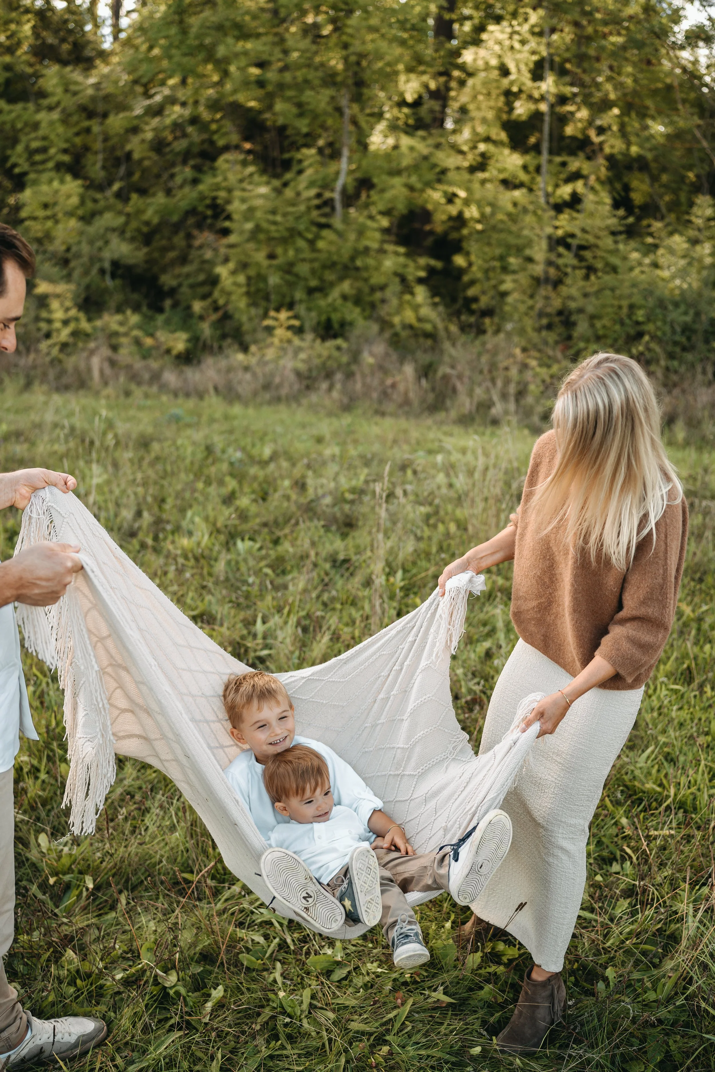 Zwei Kinder in einem Hängesessel im Grünen, von zwei Erwachsenen gehalten, im Hintergrund Bäume und Natur.