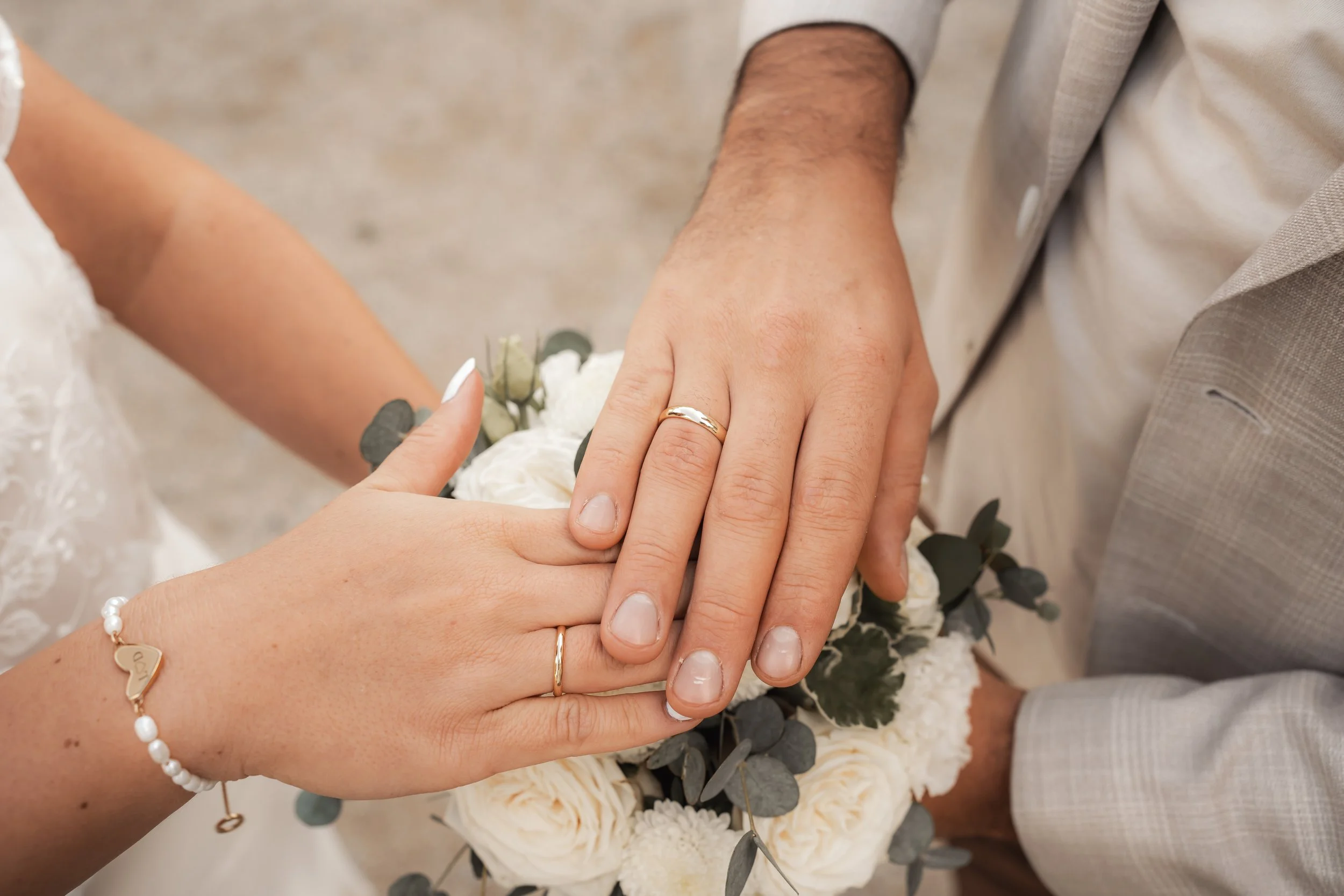 Hände eines Brautpaares mit Eheringen, über einem Brautstrauß aus weißen Blumen und grünem Laub