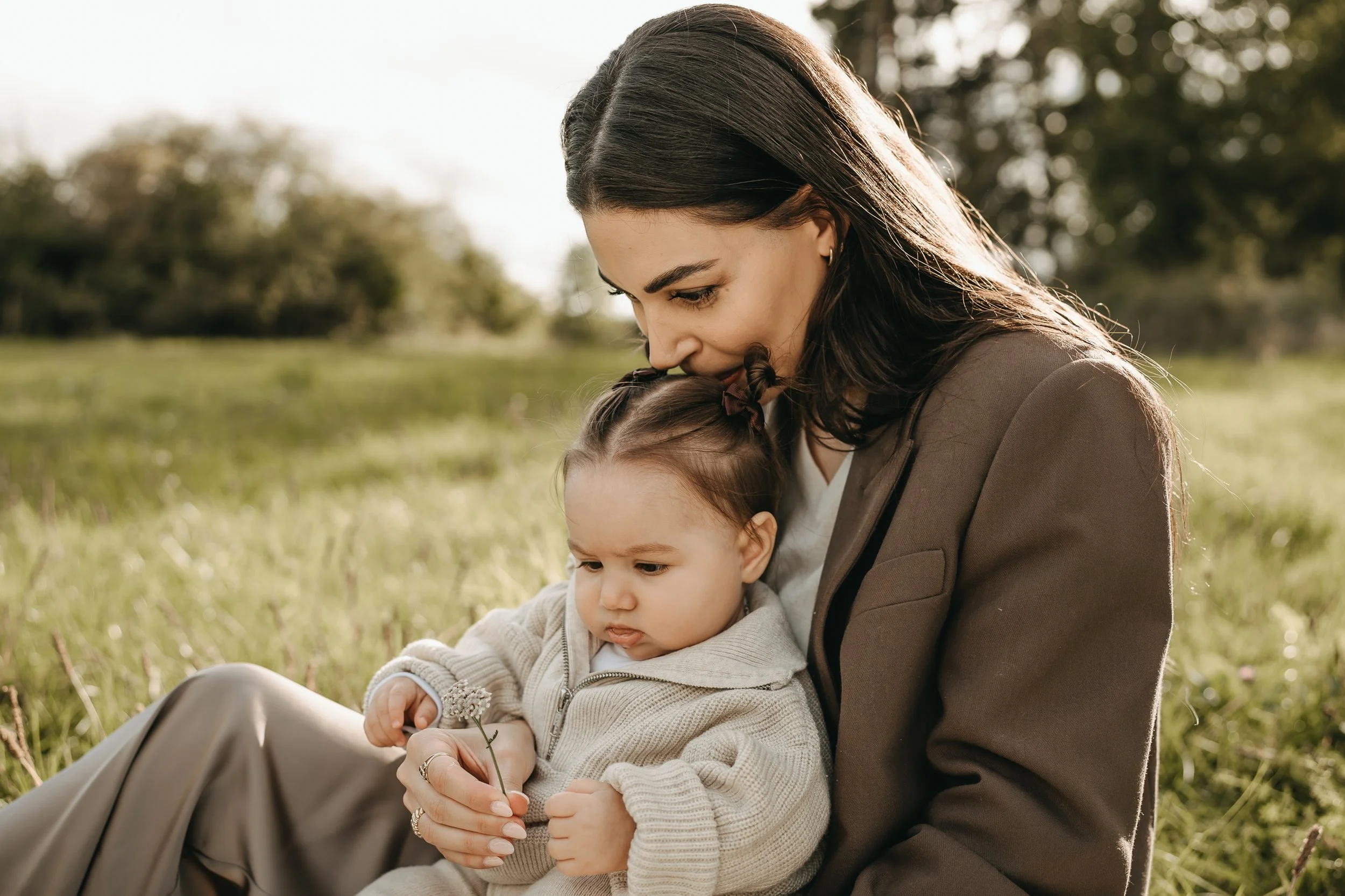 Eine Frau hält ein kleines Kind im Grünen bei Sonnenuntergang. Sie sitzen auf dem Gras, die Frau küsst das Kind auf den Kopf, das Kind betrachtet eine kleine Blume.