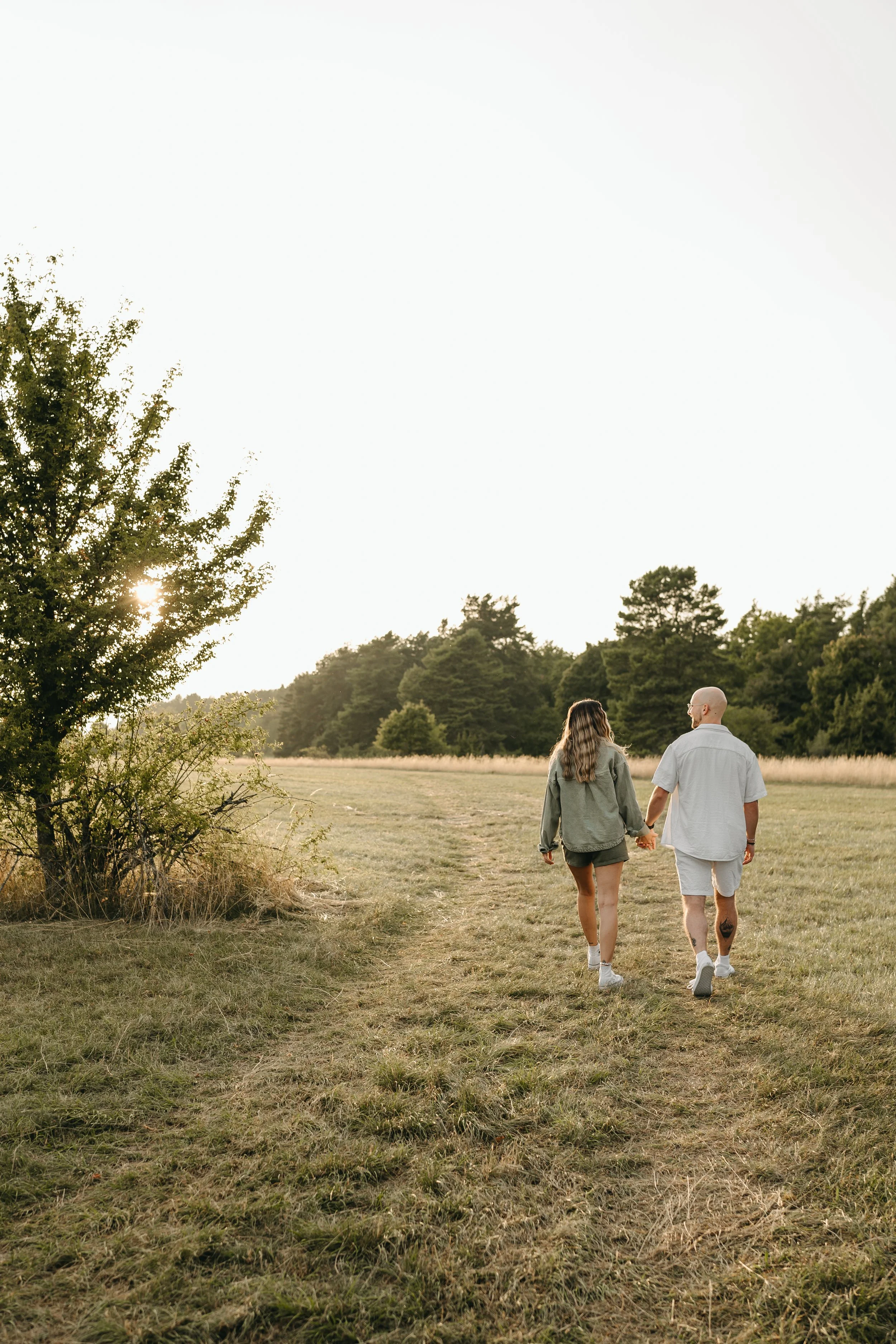 Ein Paar läuft Hand in Hand durch eine offene Wiese bei Sonnenuntergang, umgeben von Bäumen.