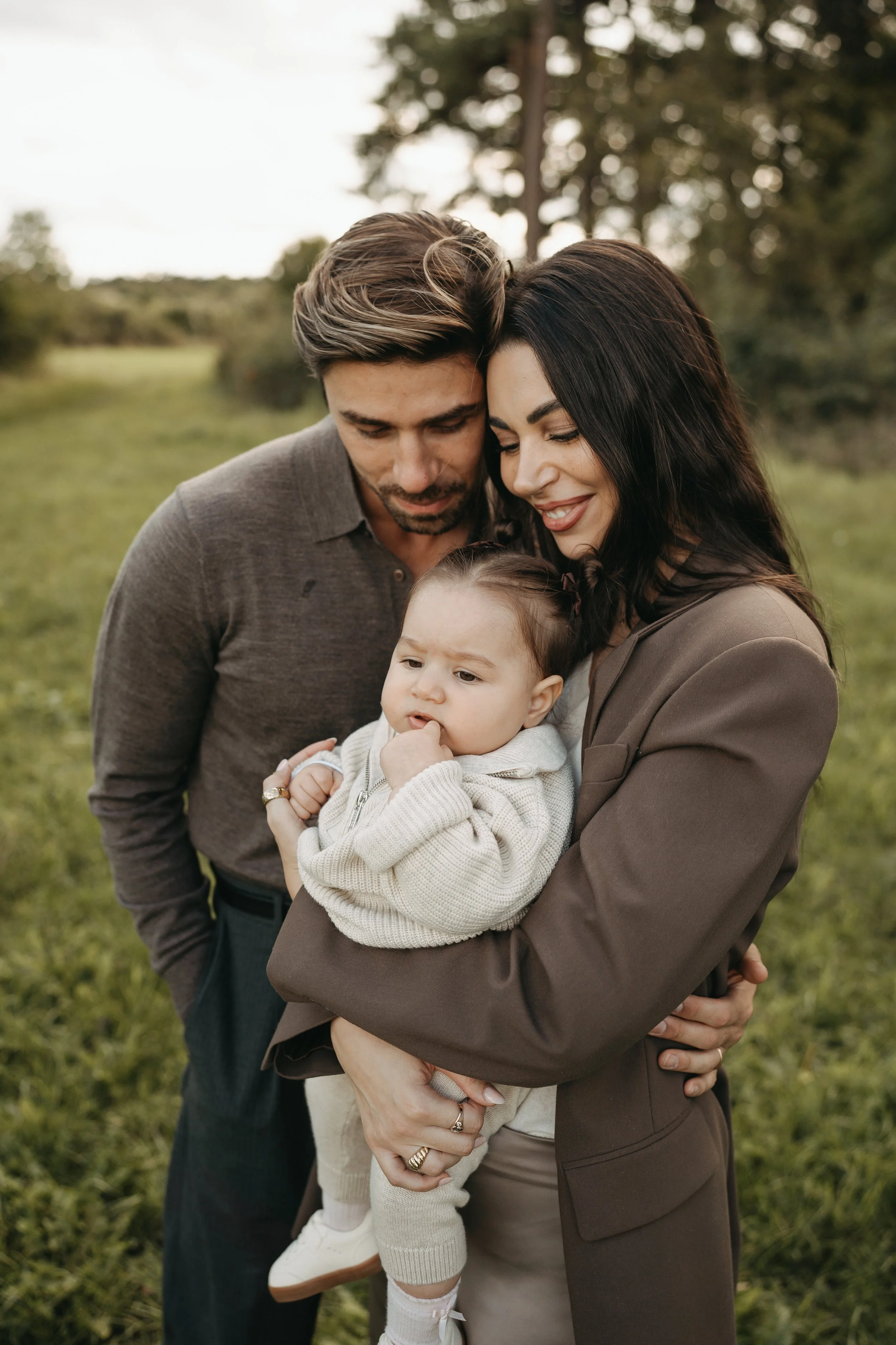 Familie mit einem kleinen Kind umarmt sich im Freien auf einer grünen Wiese mit Bäumen im Hintergrund, bei Sonnenuntergang.