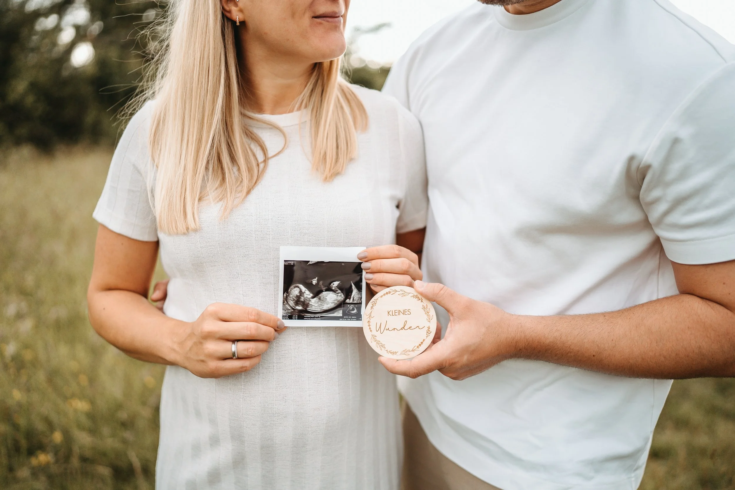 Ein Paar hält eine Ultraschallaufnahme und eine Trophäe mit der Aufschrift 'Kleines Wunder' in der Natur, vermutlich bei einer Schwangerschaftsfotografie.