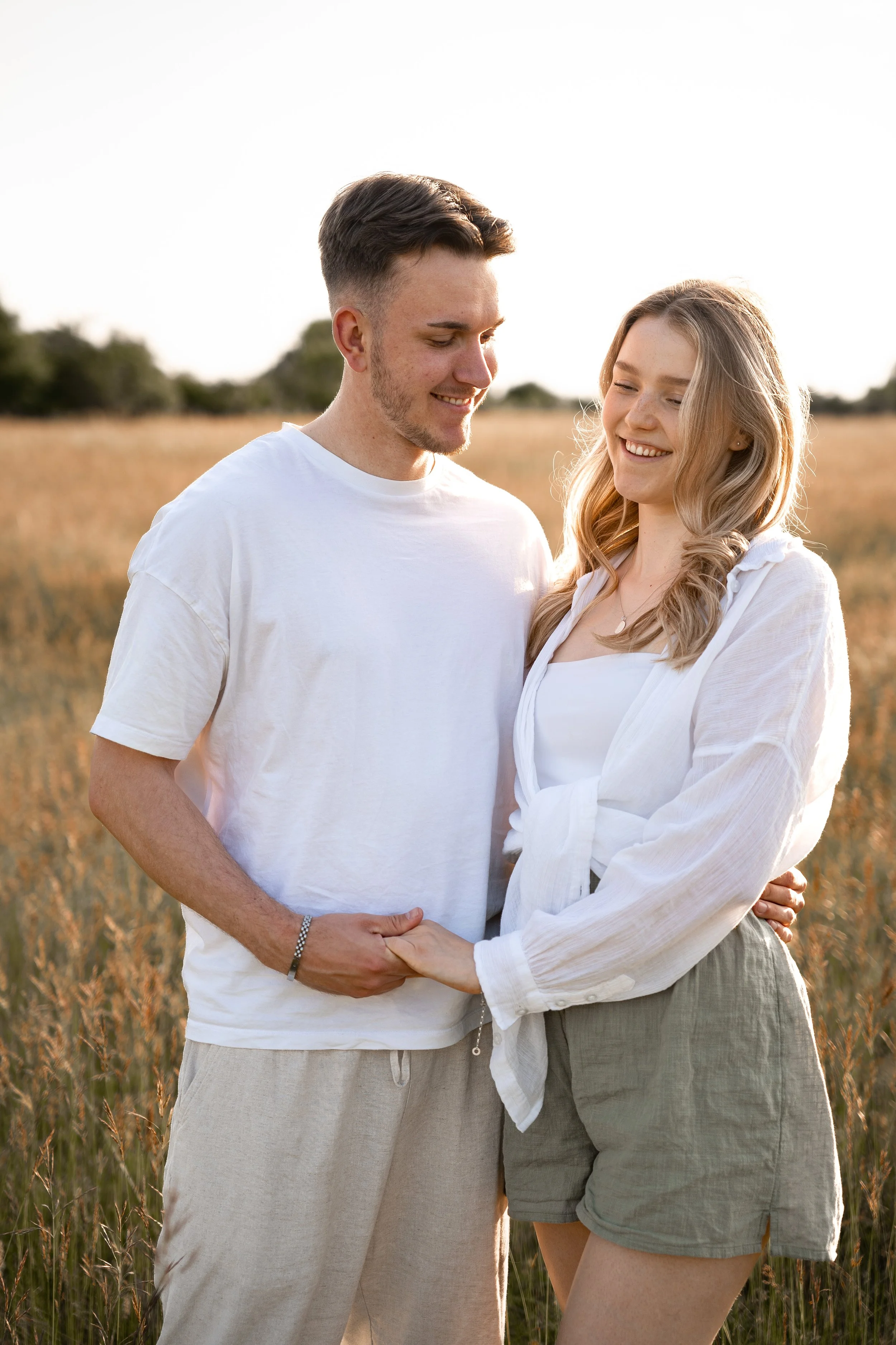 Ein lachendes Paar steht in einem goldgelben Getreidefeld bei Sonnenuntergang, Händchen haltend, in lockerer Sommerkleidung.
