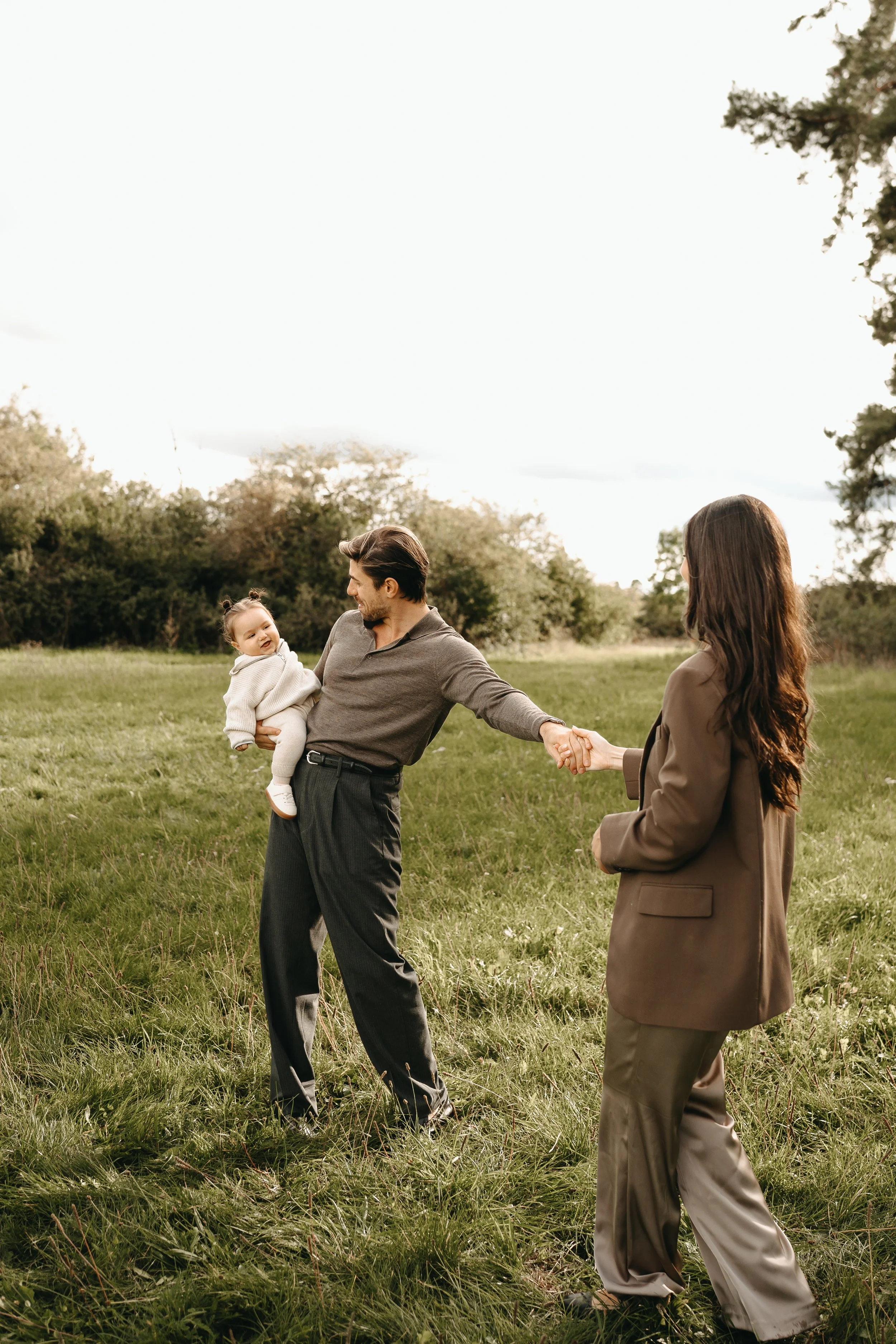 Familie im Park: Vater hält kleines Mädchen, Mutter hält Vaters Hand, im Hintergrund Bäume und Wiese.