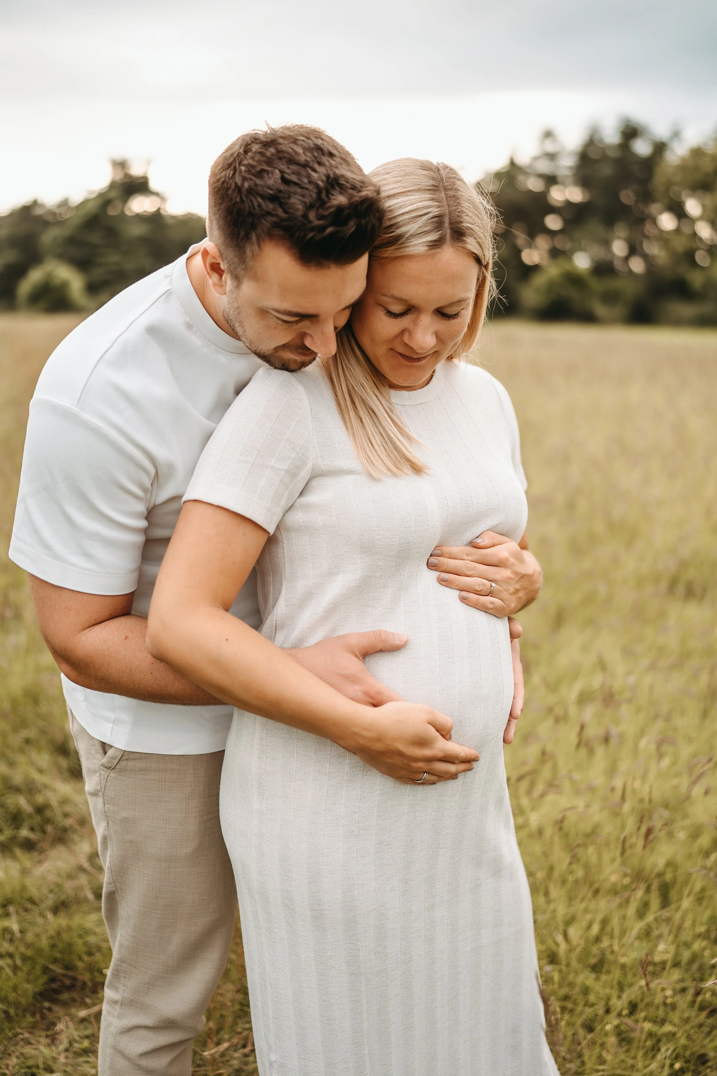 Ein Paar umarmt eine schwangere Frau in einem offenen Feld, während sie liebevoll ihren Babybauch hält.