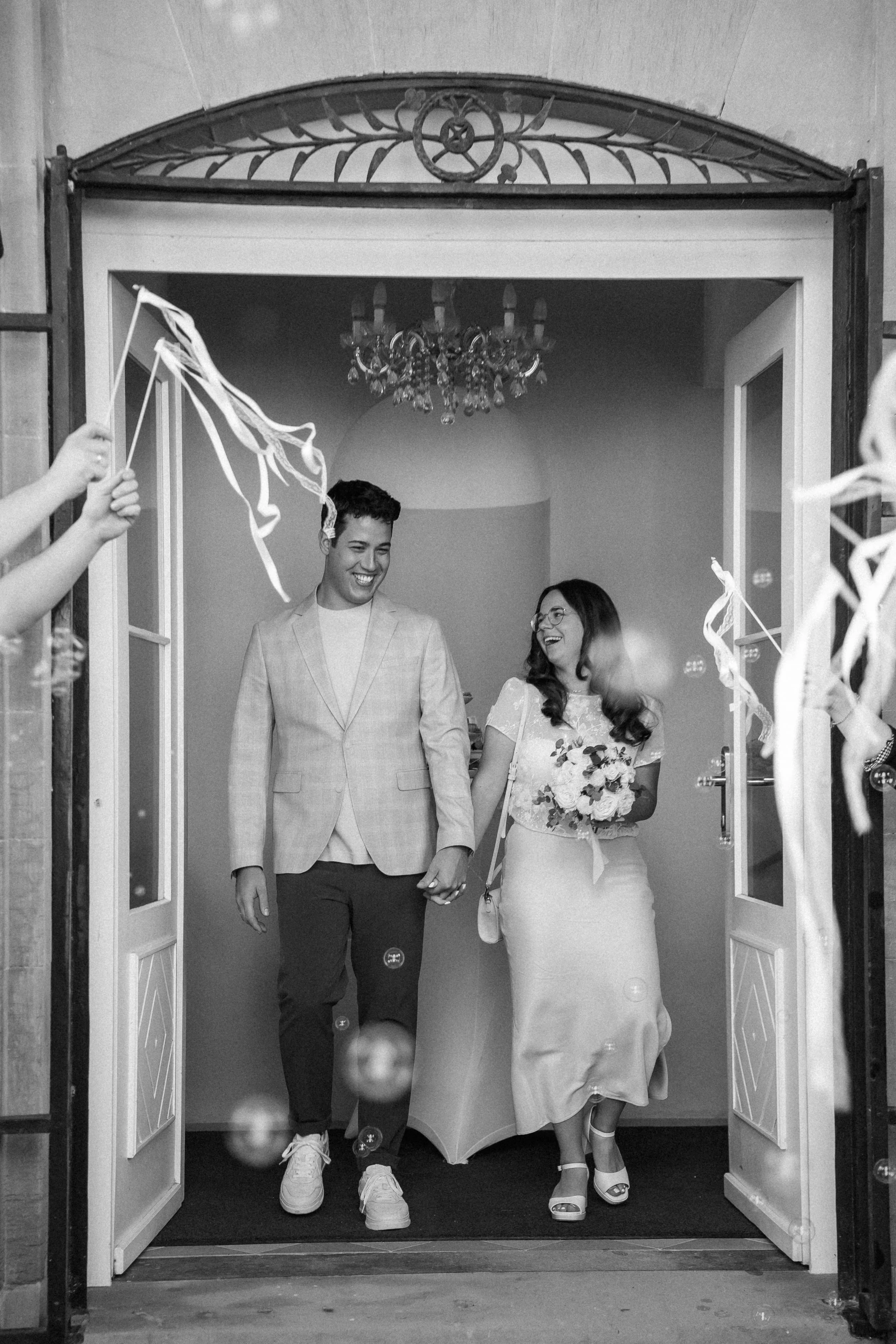 Ein glückliches Paar, wahrscheinlich Braut und Bräutigam, betritt gemeinsam einen Raum, während Gäste mit Luftballons und Seidenband jubeln.