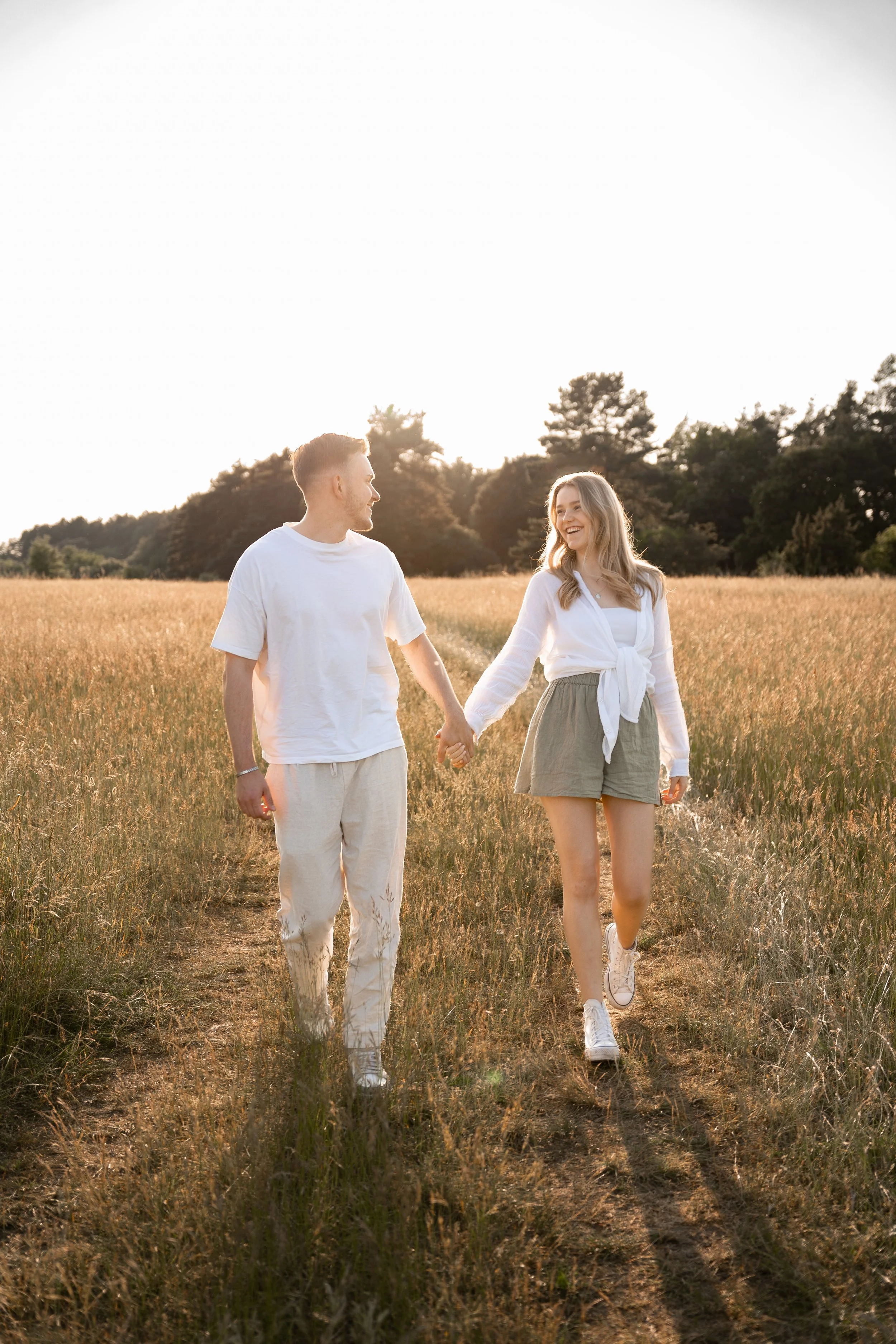 Ein Paar läuft Hand in Hand durch ein goldgelbes Feld bei Sonnenuntergang und lächelt sich an.
