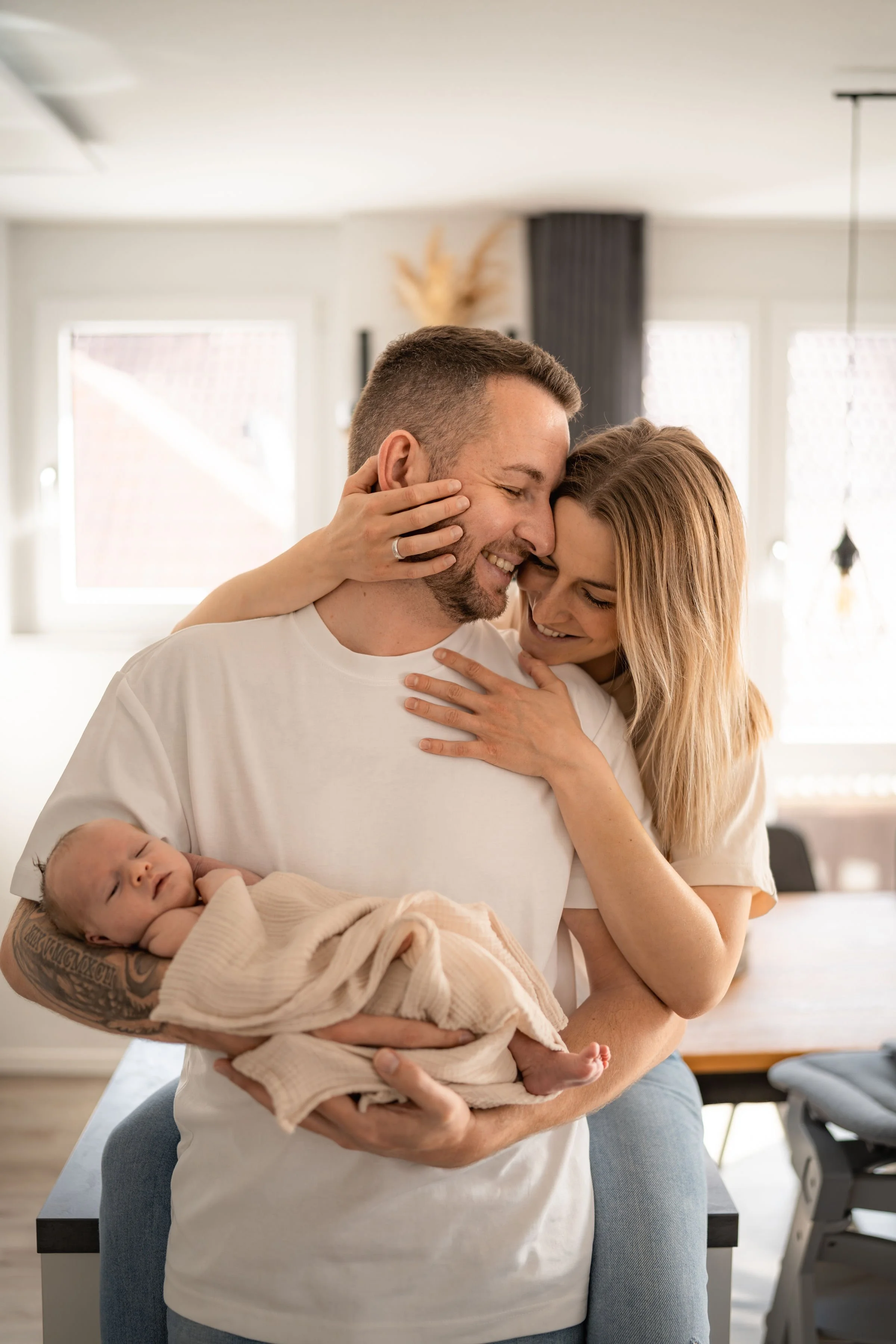 Ein glückliches Paar hält ein schlafendes Baby in den Armen, umgeben von natürlichem Licht in einer modernen Wohnung.
