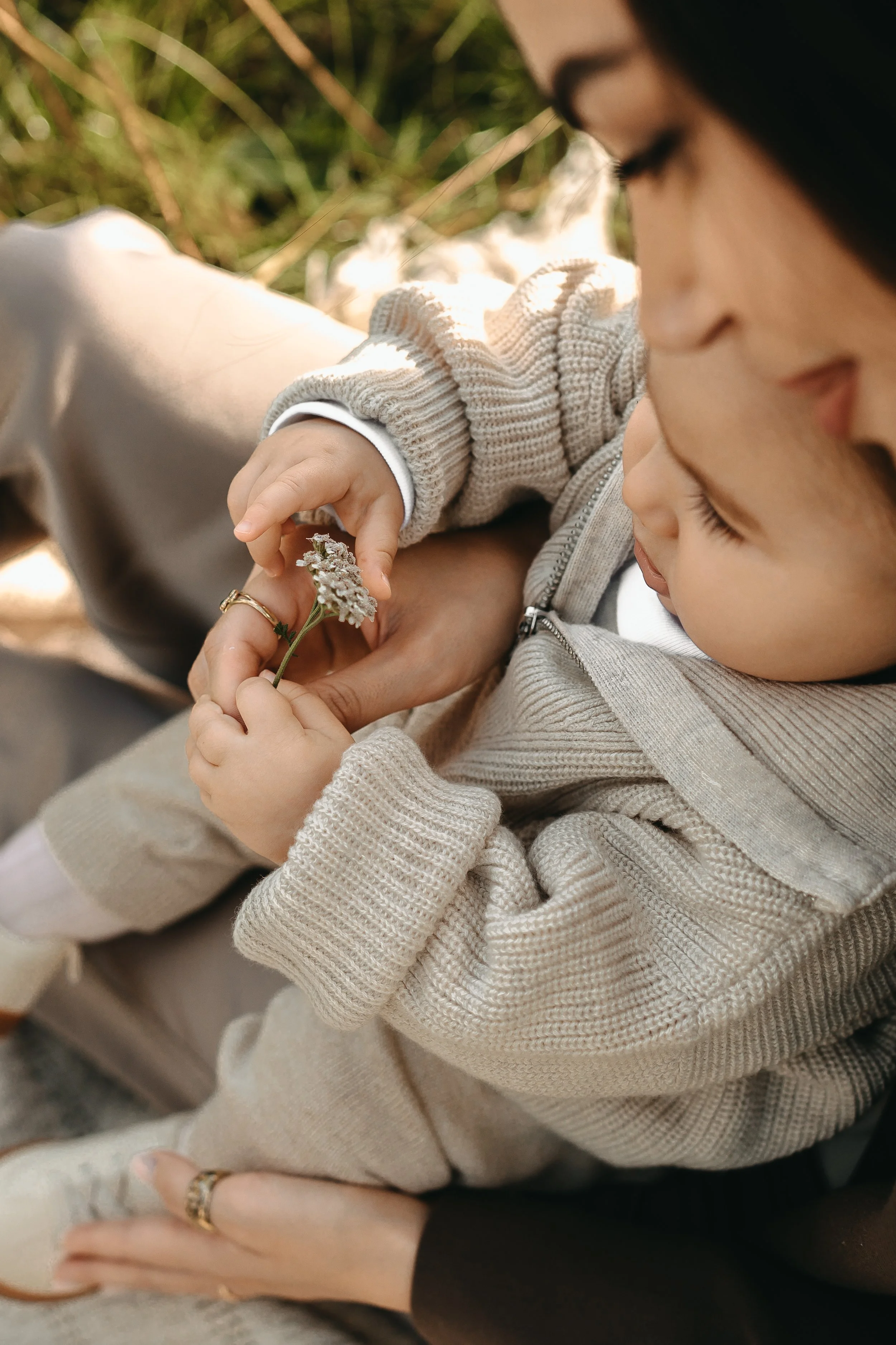 Ein Kind hält eine kleine Blume und zeigt sie einer erwachsenen Frau, die in einer natürlichen Umgebung sitzt, vermutlich im Freien.